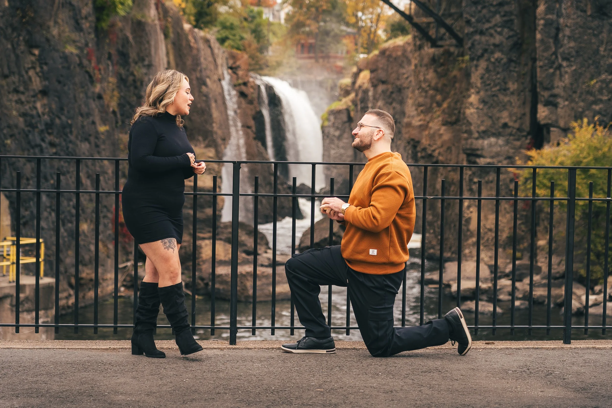 A man proposing marriage to a woman by a waterfall, with the man kneeling and the woman standing, both smiling, in a scenic outdoor setting.