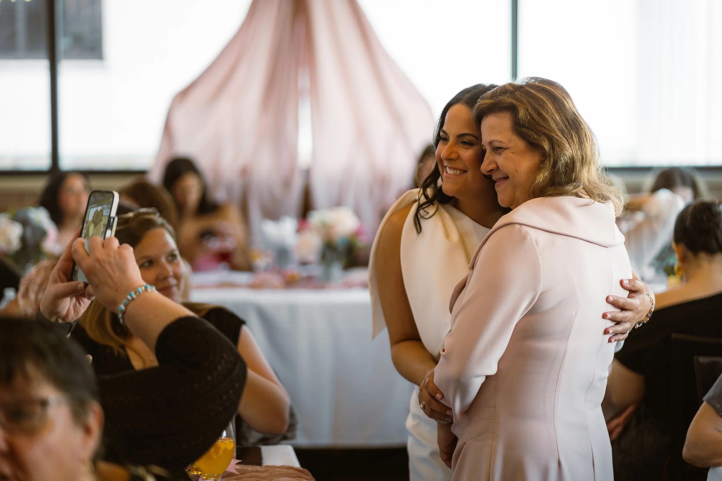 Two women are hugging at a social event, smiling and taking a photo together while others sit at tables in the background.