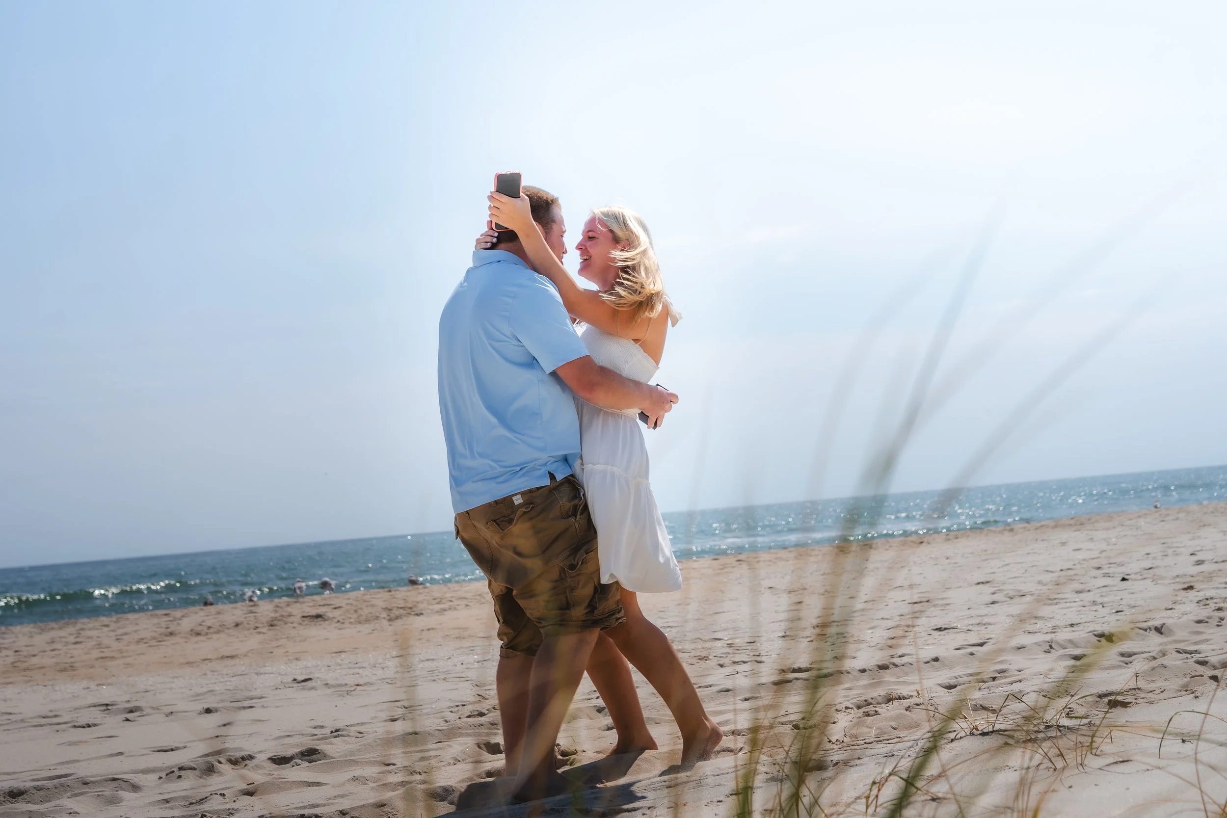A couple dancing on the beach, with the man holding a phone while they smile at each other under a bright sky.