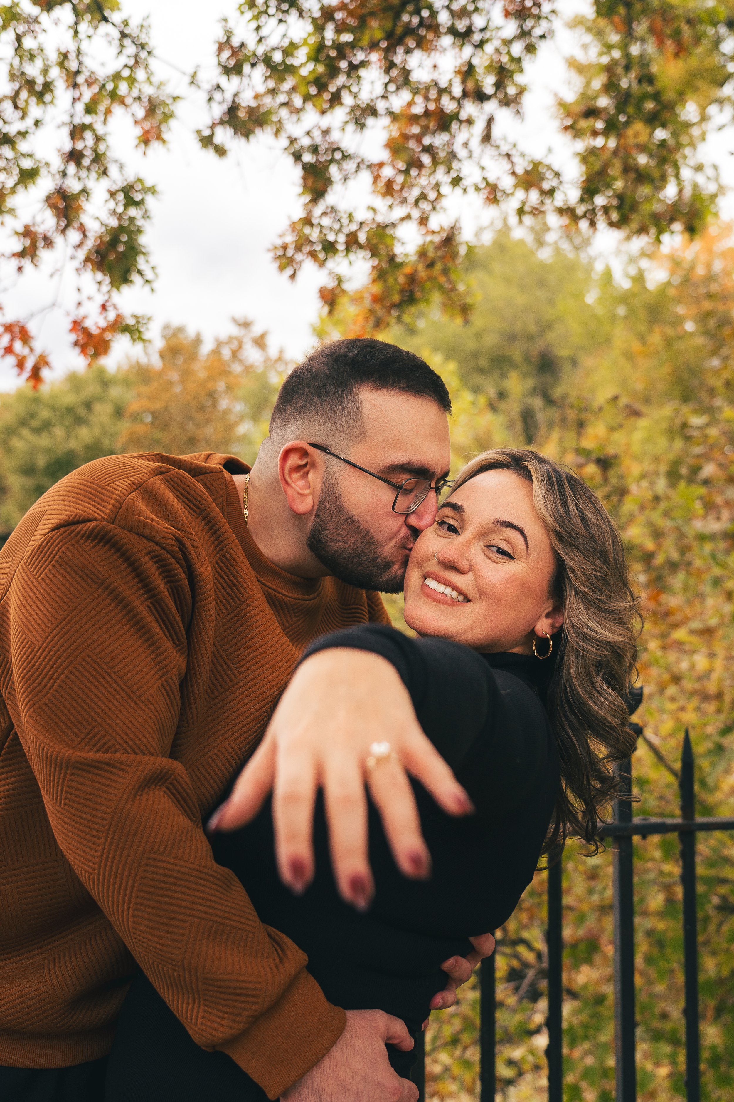 A couple embracing outdoors during fall, with the man kissing the woman on her cheek as she smiles, surrounded by autumnal trees with colorful leaves.