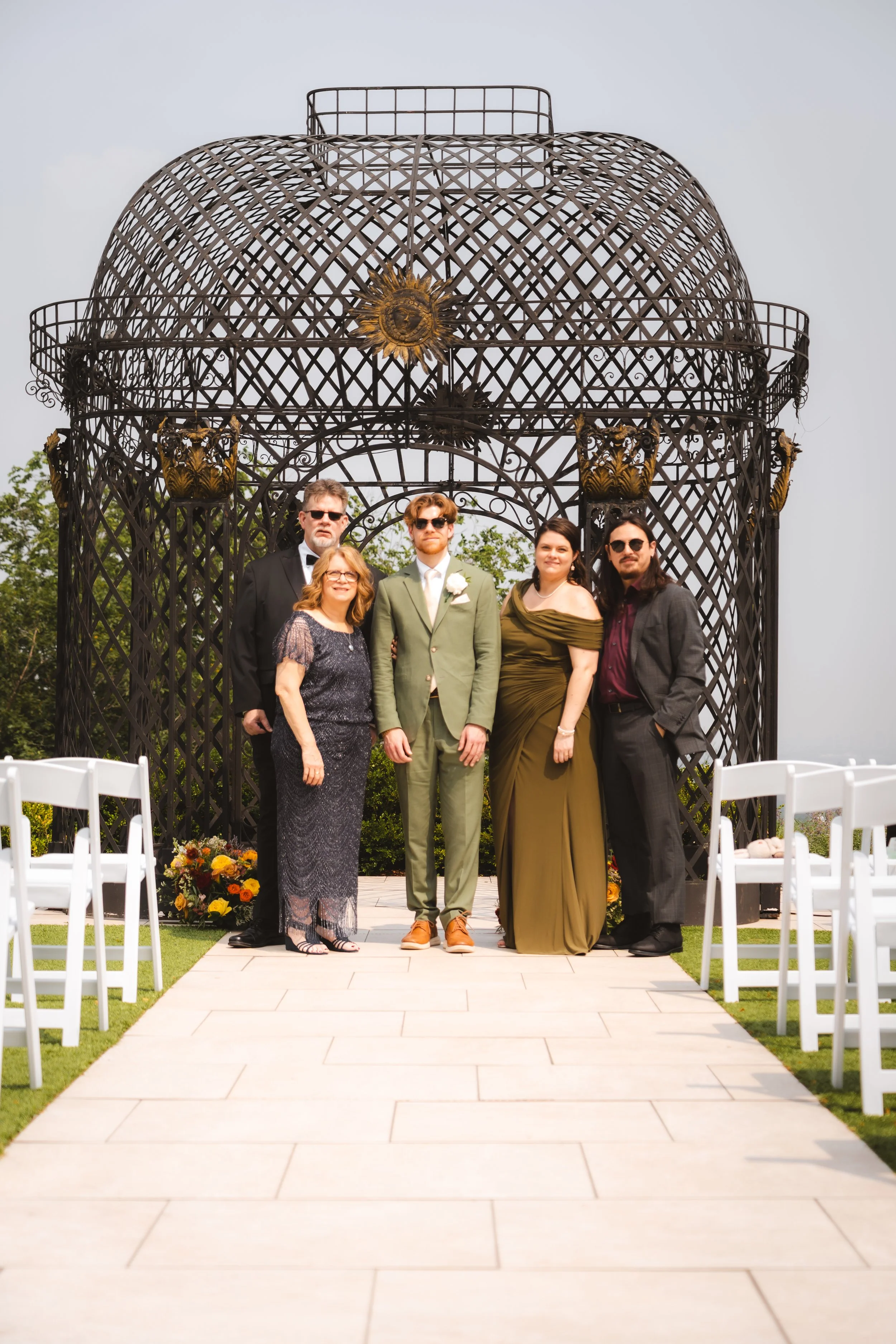A wedding ceremony with five people standing on an outdoor aisle in front of a decorative metal gazebo, with white chairs on each side and a flower arrangement at the base of the gazebo.