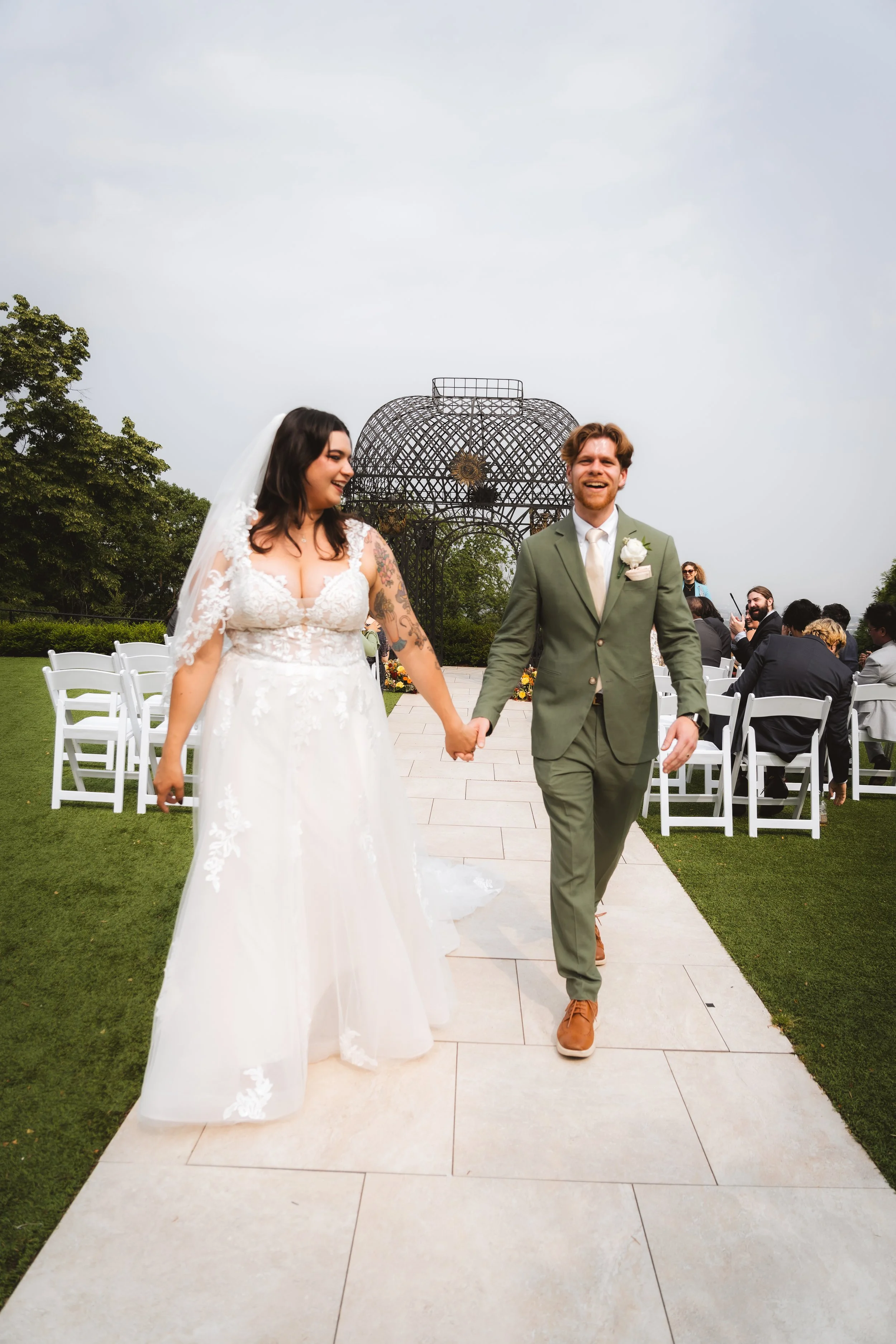 A bride and groom walking hand in hand on a wedding aisle outdoors, smiling, with wedding guests seated on white chairs in the background and a decorative metal structure behind them.
