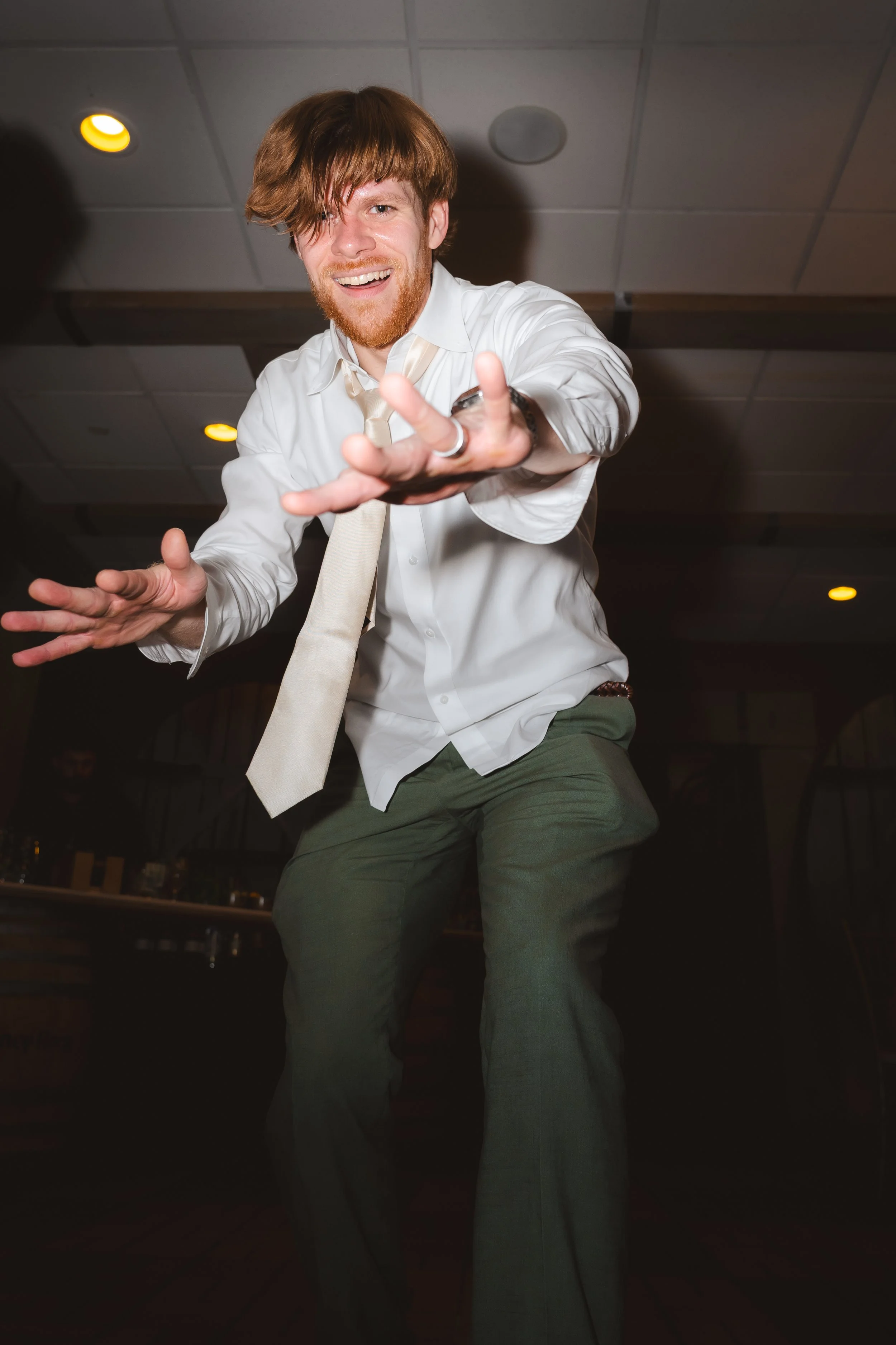 A young man with red hair and a beard is dancing and smiling in a dimly lit indoor setting, wearing a white shirt, a cream-colored tie, and green pants.