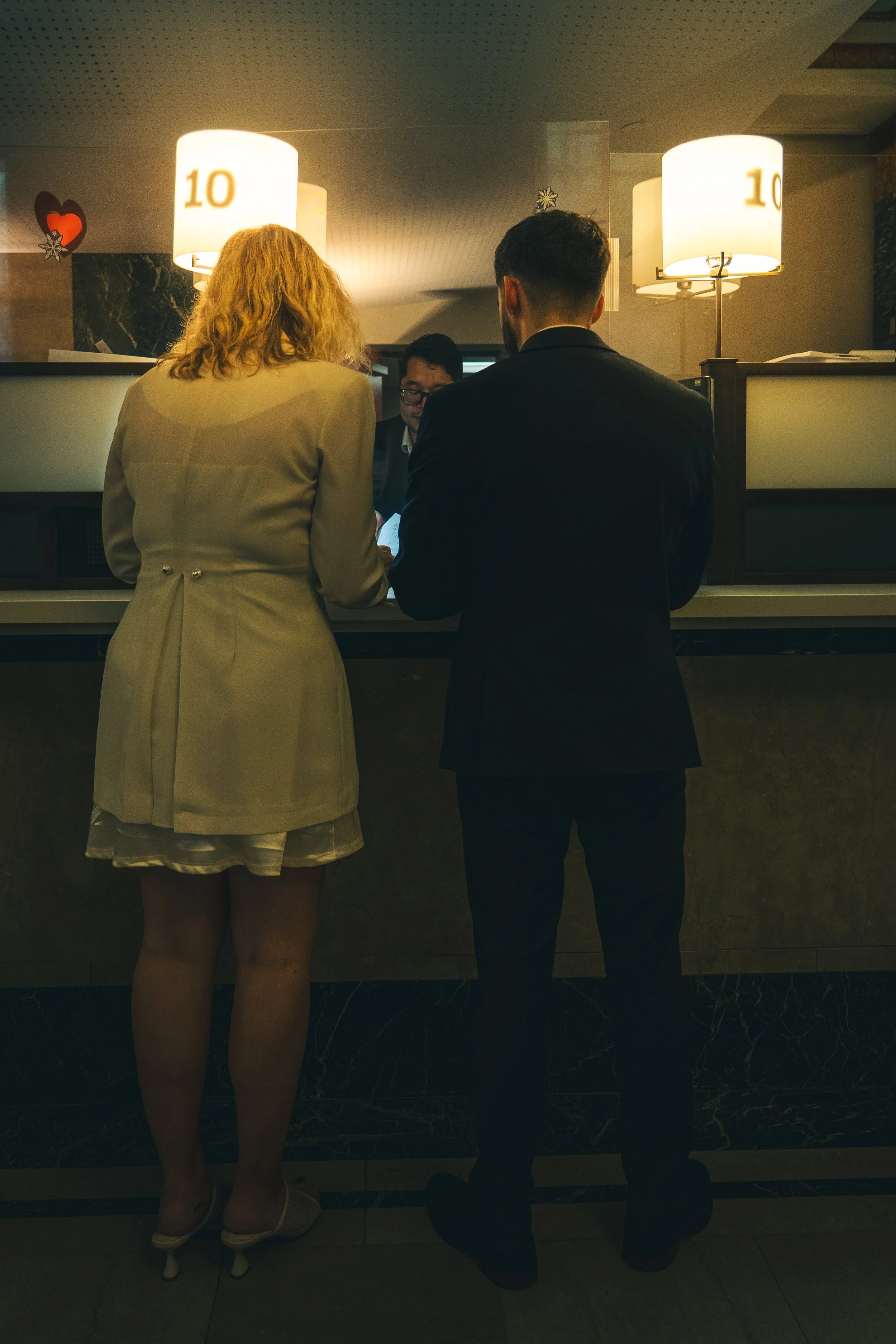 A man and woman standing at a hotel reception desk, looking at a tablet held by a receptionist. The woman has blonde hair and is wearing a beige coat and dress, while the man has dark hair and a beard, dressed in a dark suit. The reception area has m