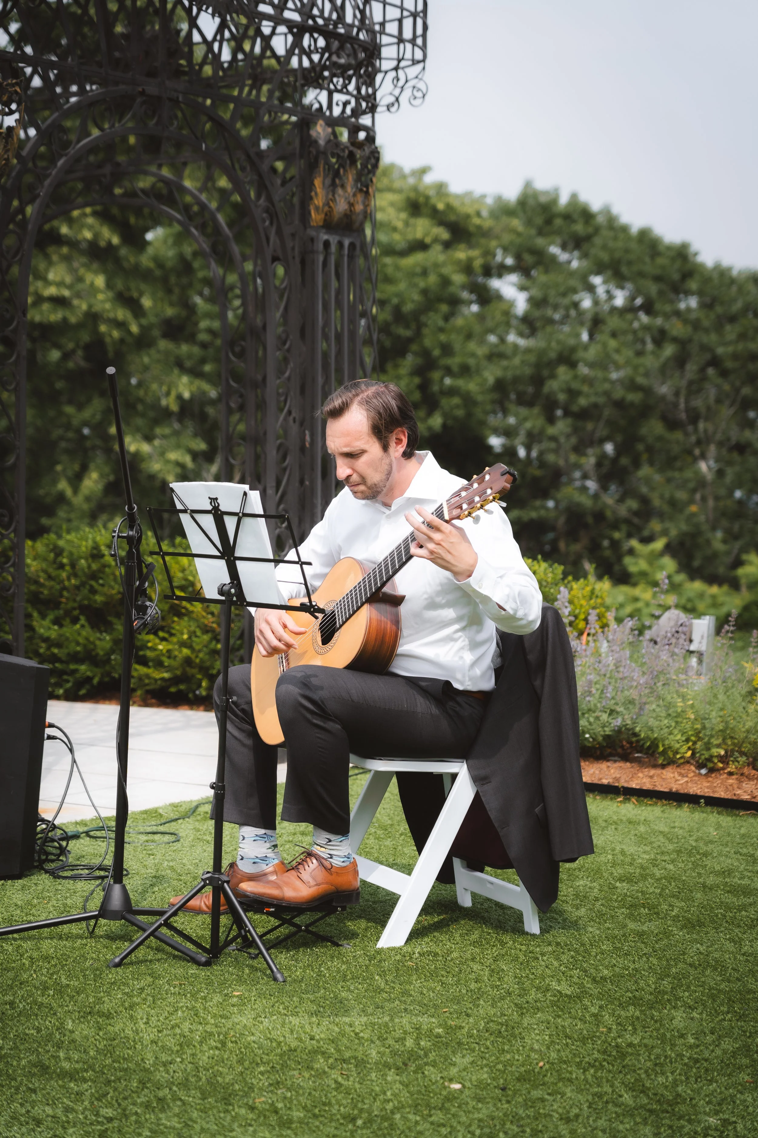 A man in a white shirt and black pants playing an acoustic guitar outdoors during the daytime, sitting on a white chair with a music stand and microphone nearby, with trees and greenery in the background.