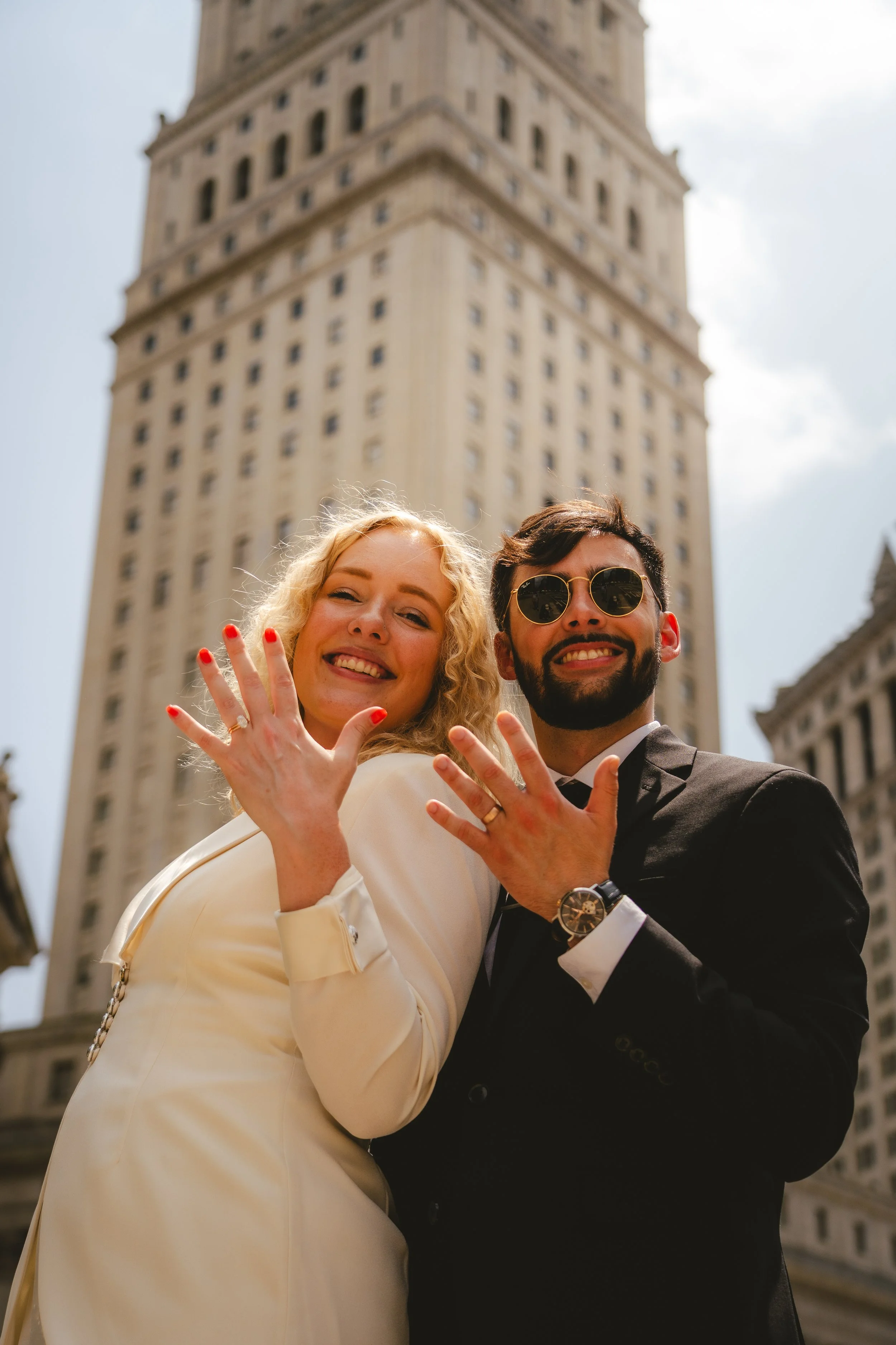 A smiling couple, a woman with curly blonde hair in a white dress and a man with dark hair and sunglasses in a black suit, showing their wedding rings, standing in front of a tall building.