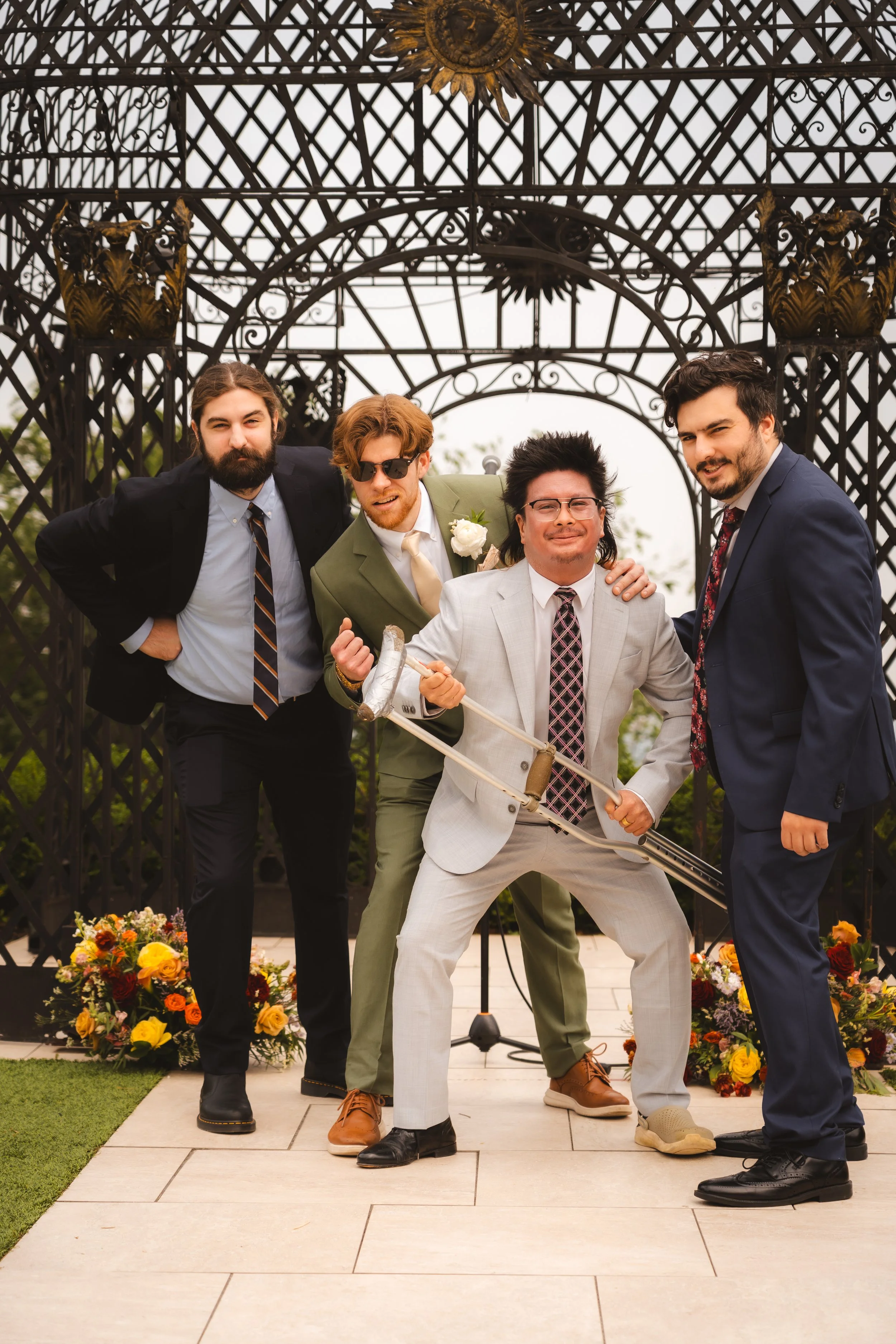 Four men dressed in suits, posing playfully outdoors with a decorative metal archway in the background and flower arrangements on the ground.