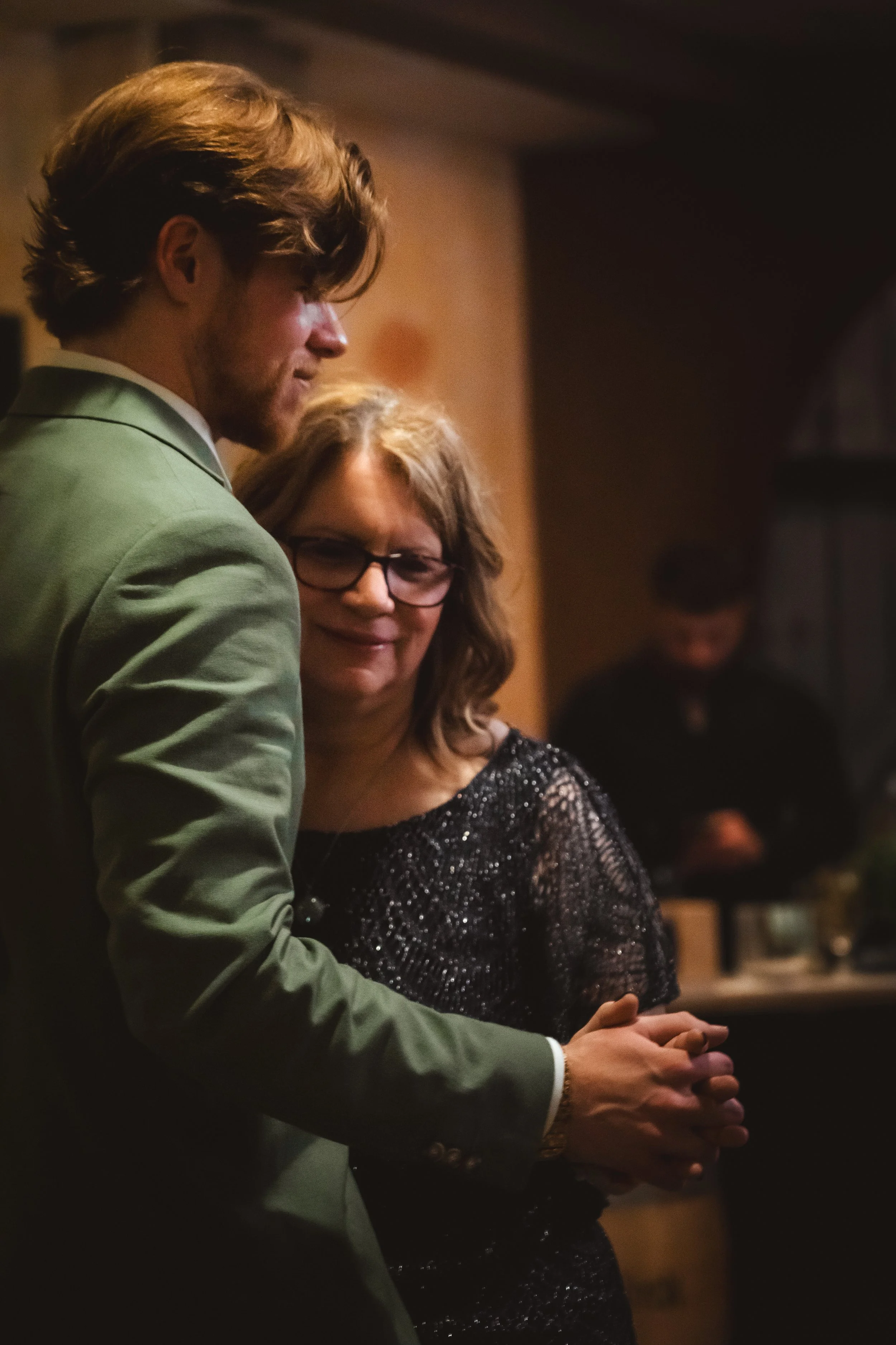 A young man in a green suit and a woman with glasses and a black sparkly dress dancing and holding hands at an indoor event.
