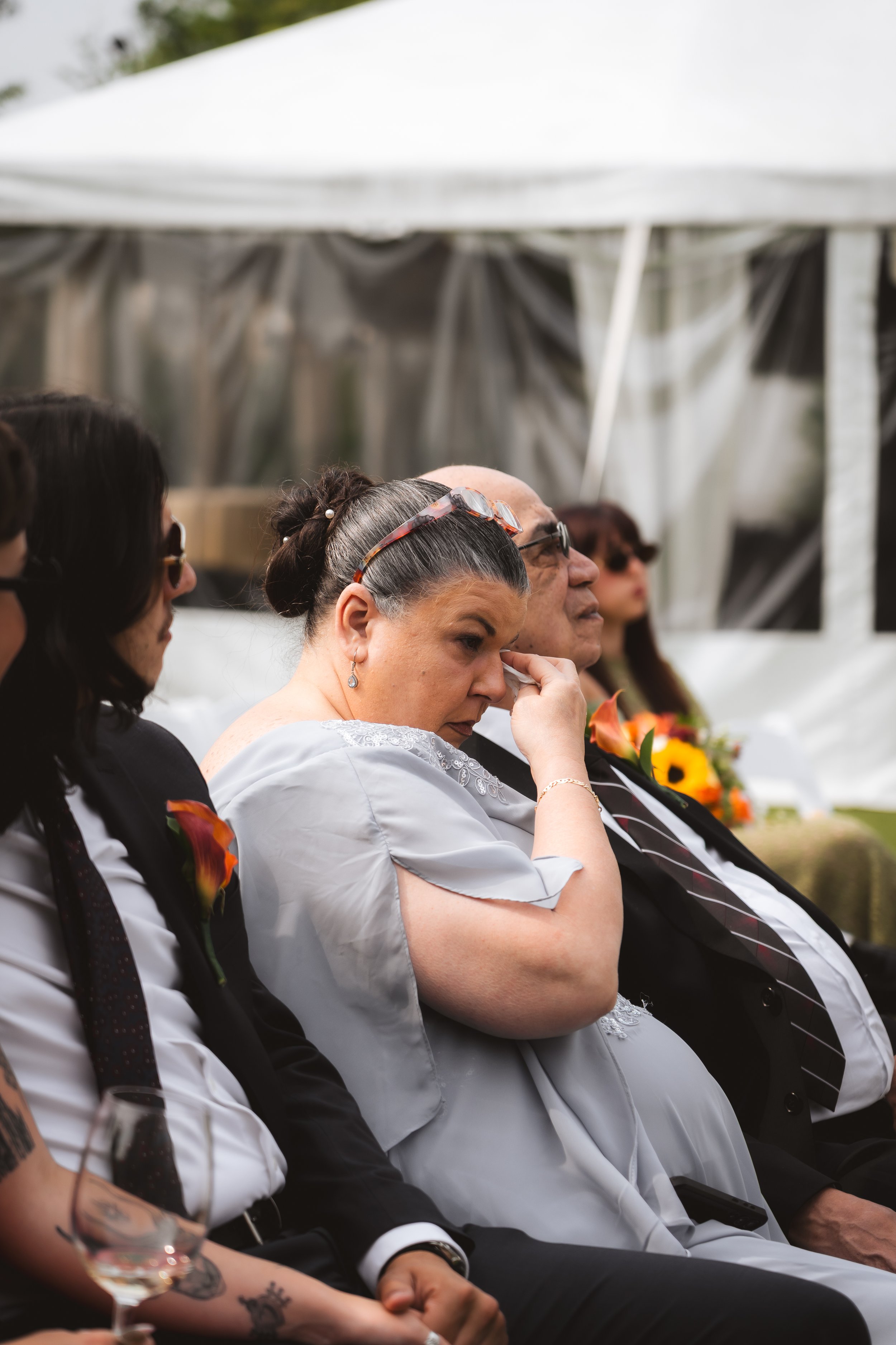 Group of adults seated outdoors at a formal event, with a white canopy tent in the background. One woman is wiping a tear from her eye, suggesting an emotional moment.