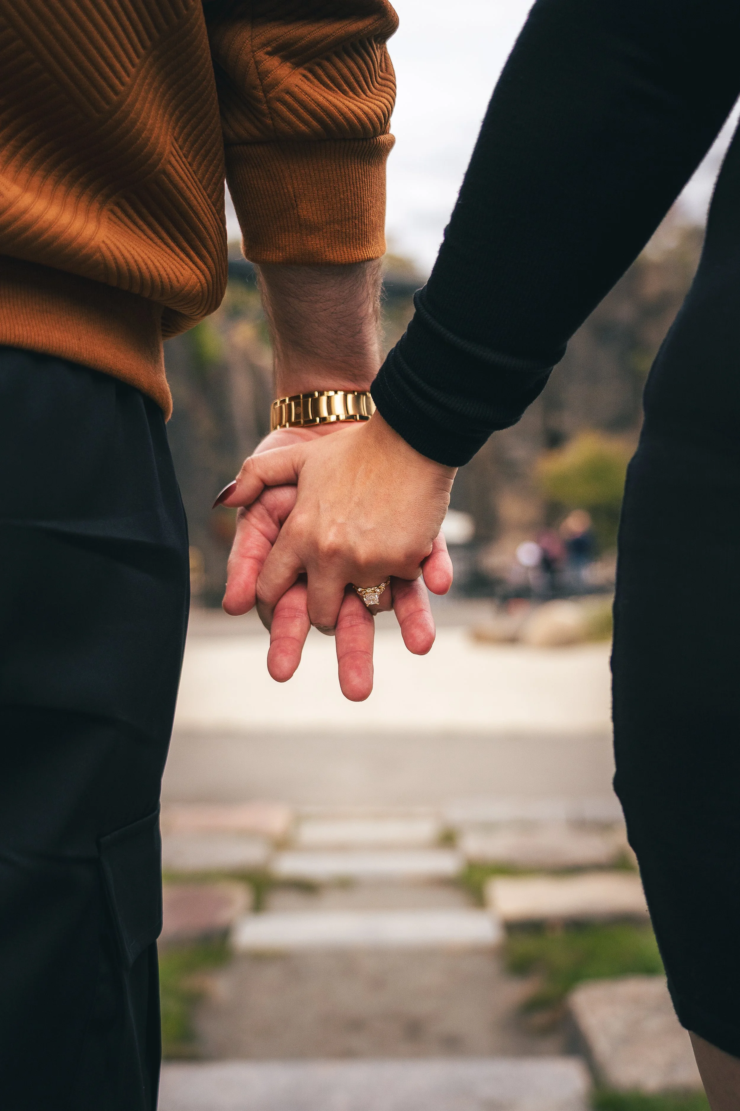 Close-up of a couple holding hands outdoors, with blurred background of park and pathway.