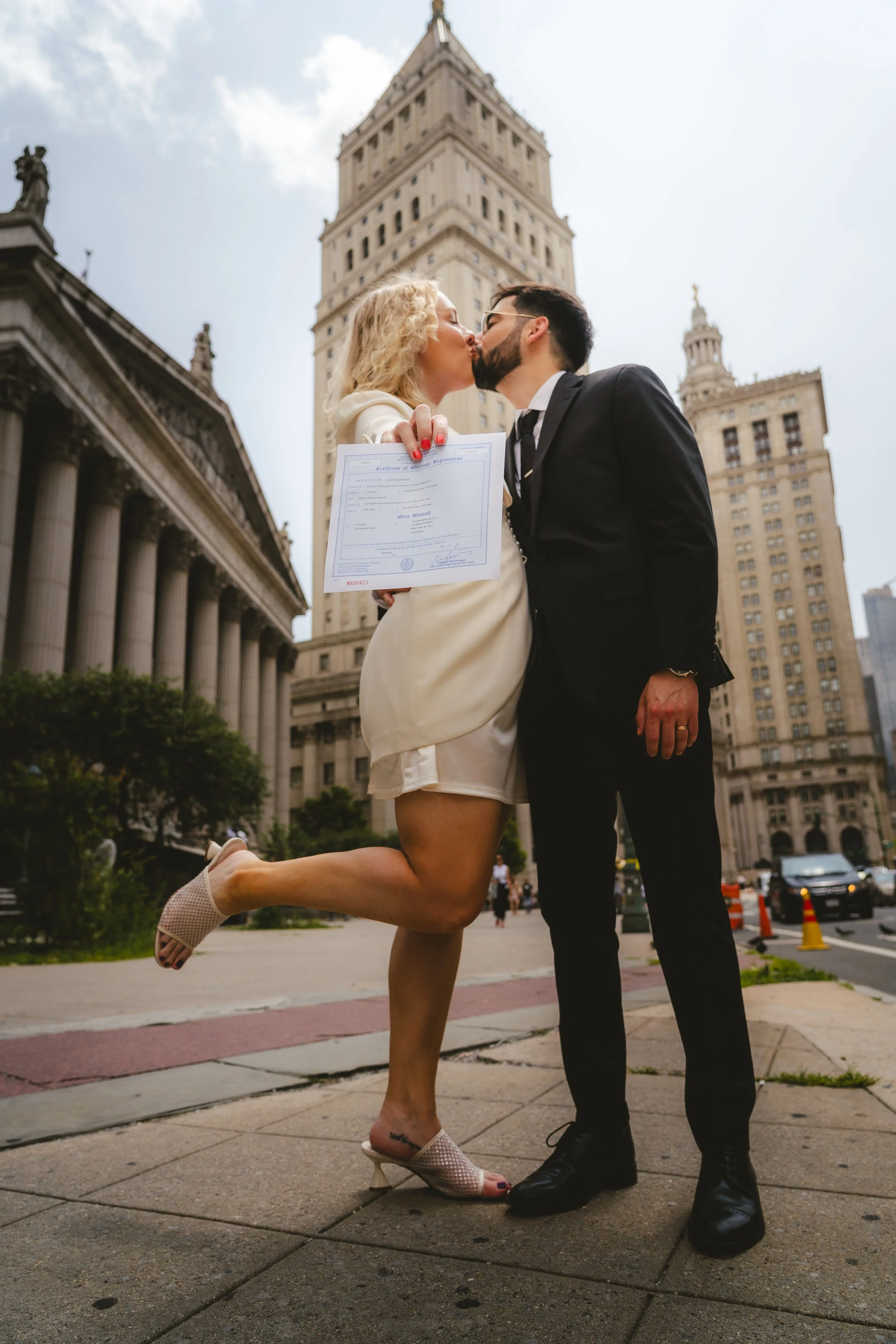 A couple in formal attire sharing a kiss in front of a tall city building, with the woman holding a marriage certificate.