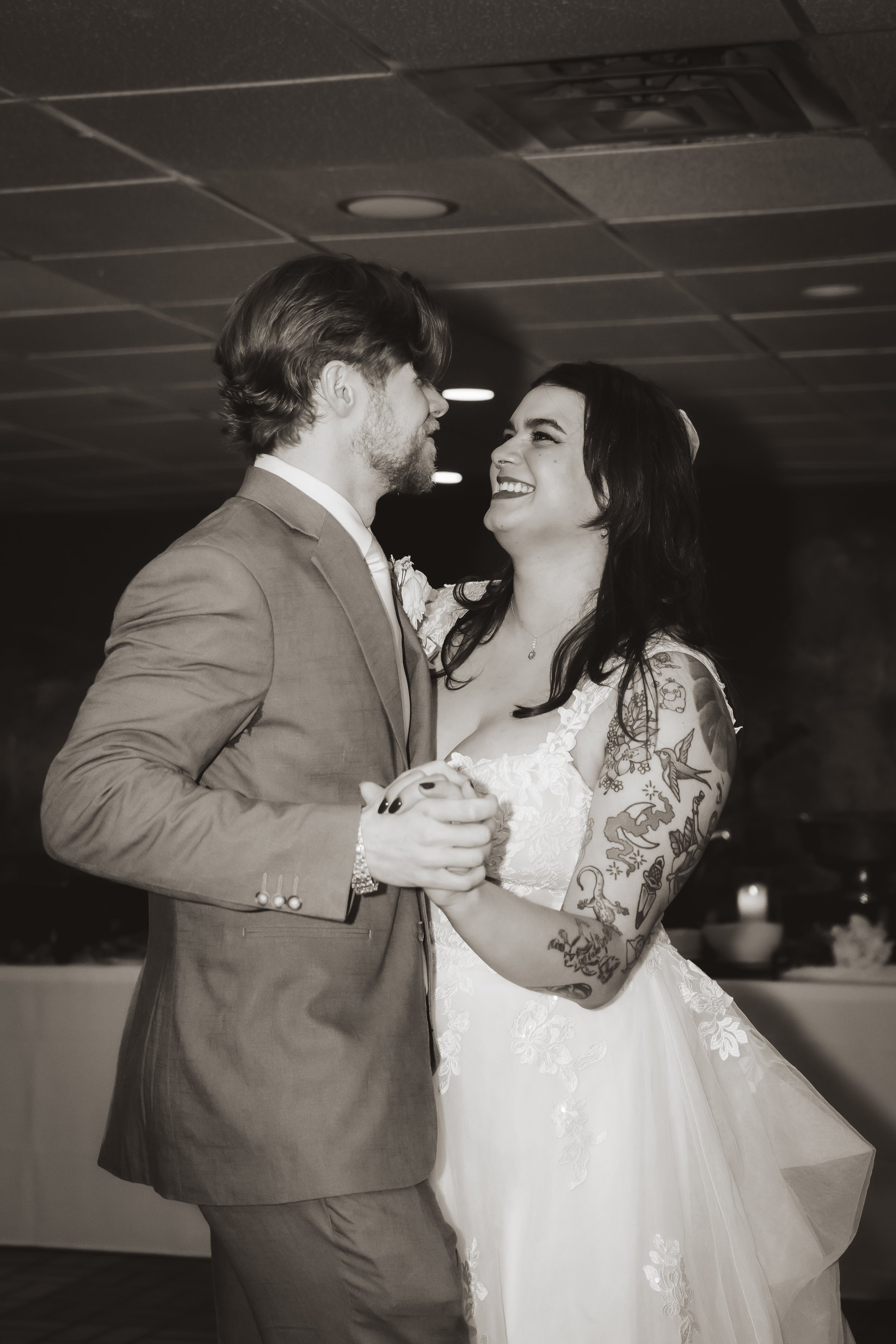 A newlywed couple dancing, the groom in a suit and the bride in a lace wedding dress with tattoos on her arms, smiling at each other.