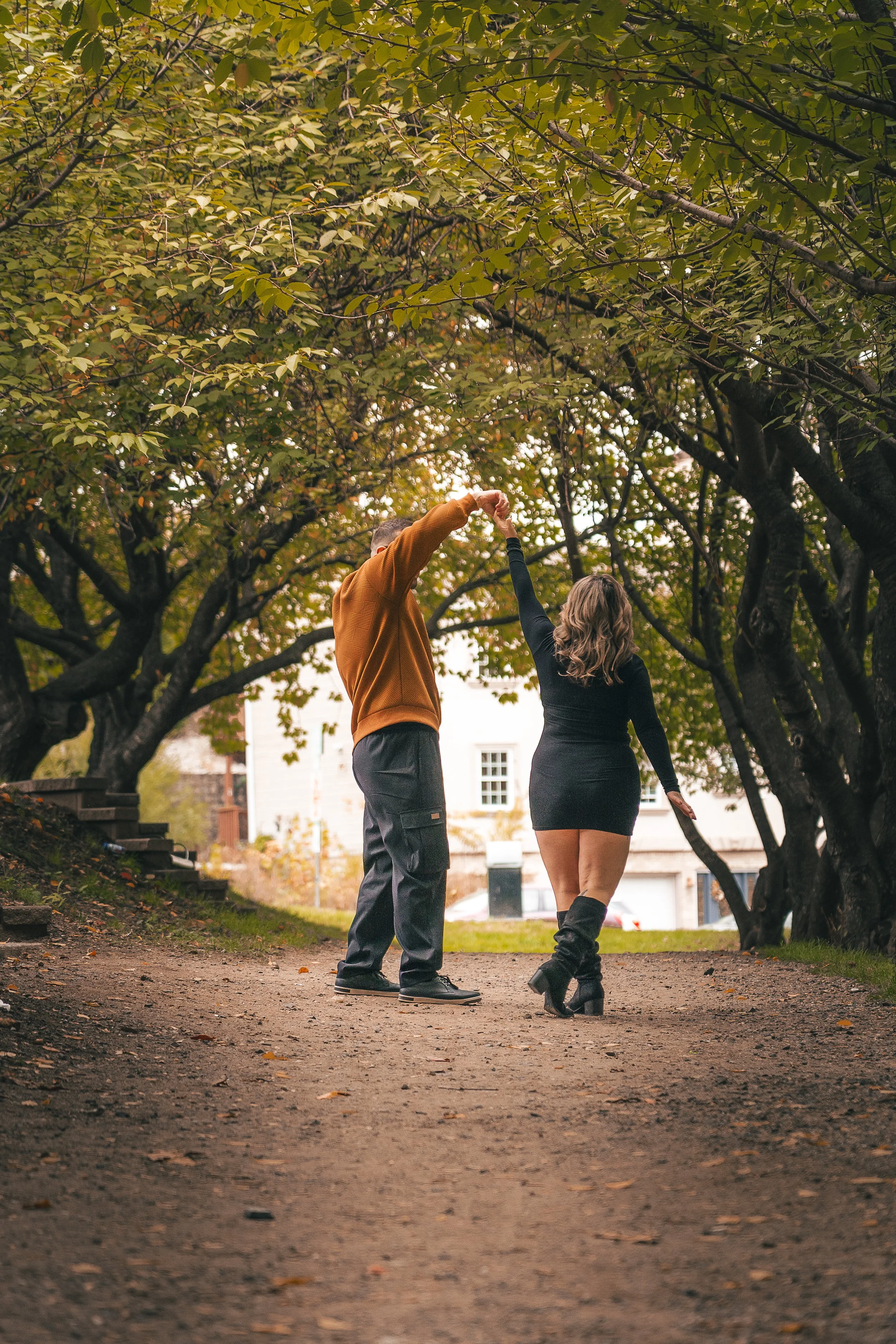 A couple dancing under trees in a park with green foliage and a residential building in the background.