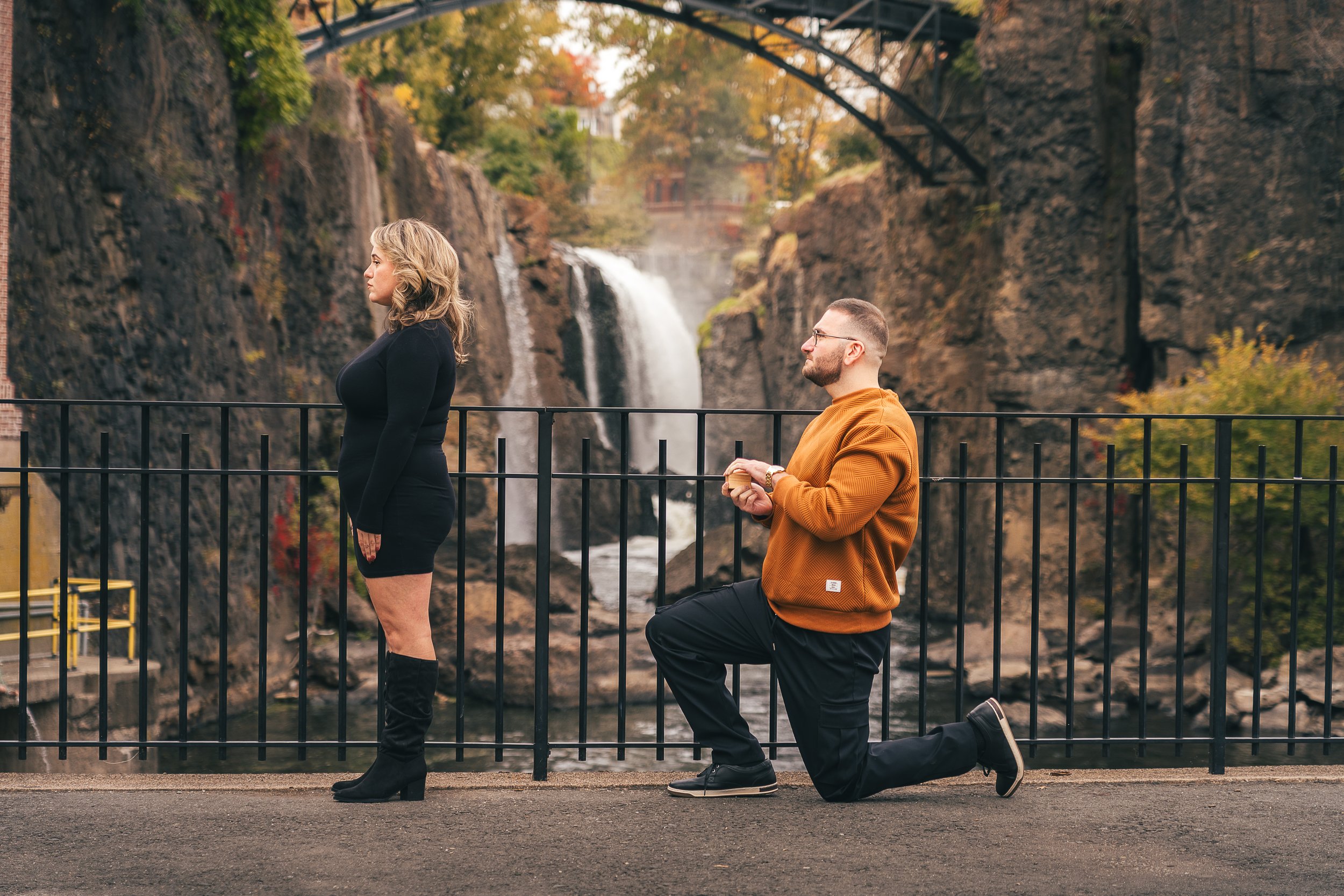 A man kneeling on one knee proposing to a woman standing in front of a black metal fence with a waterfall and rocky cliffs in the background.