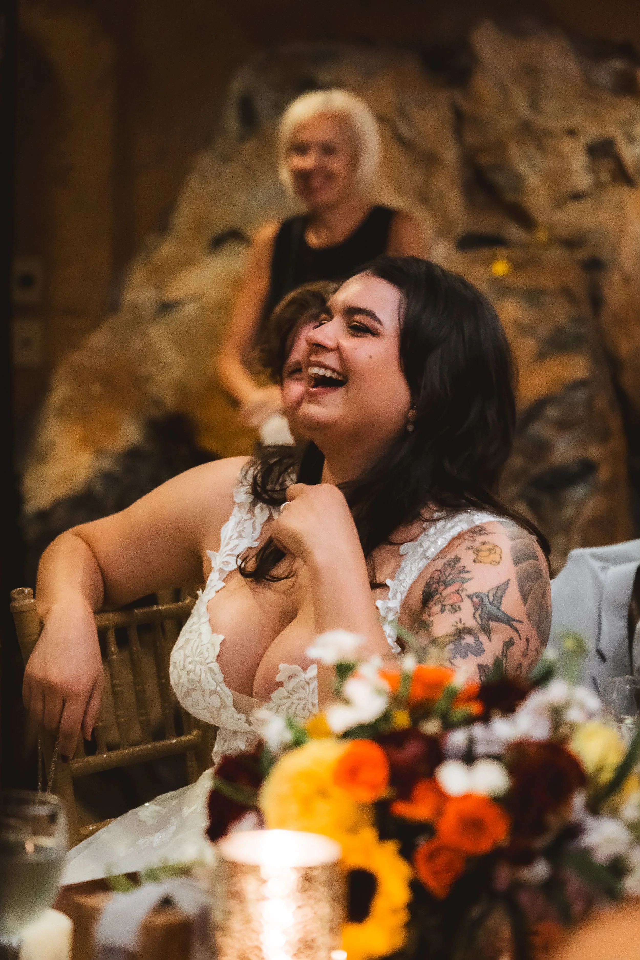 A woman with black hair and tattoos on her arm, wearing a white dress, is laughing at a celebration or gathering, with a floral arrangement and candle in the foreground, and a woman with blonde hair in the background, all indoors with a rocky wall.