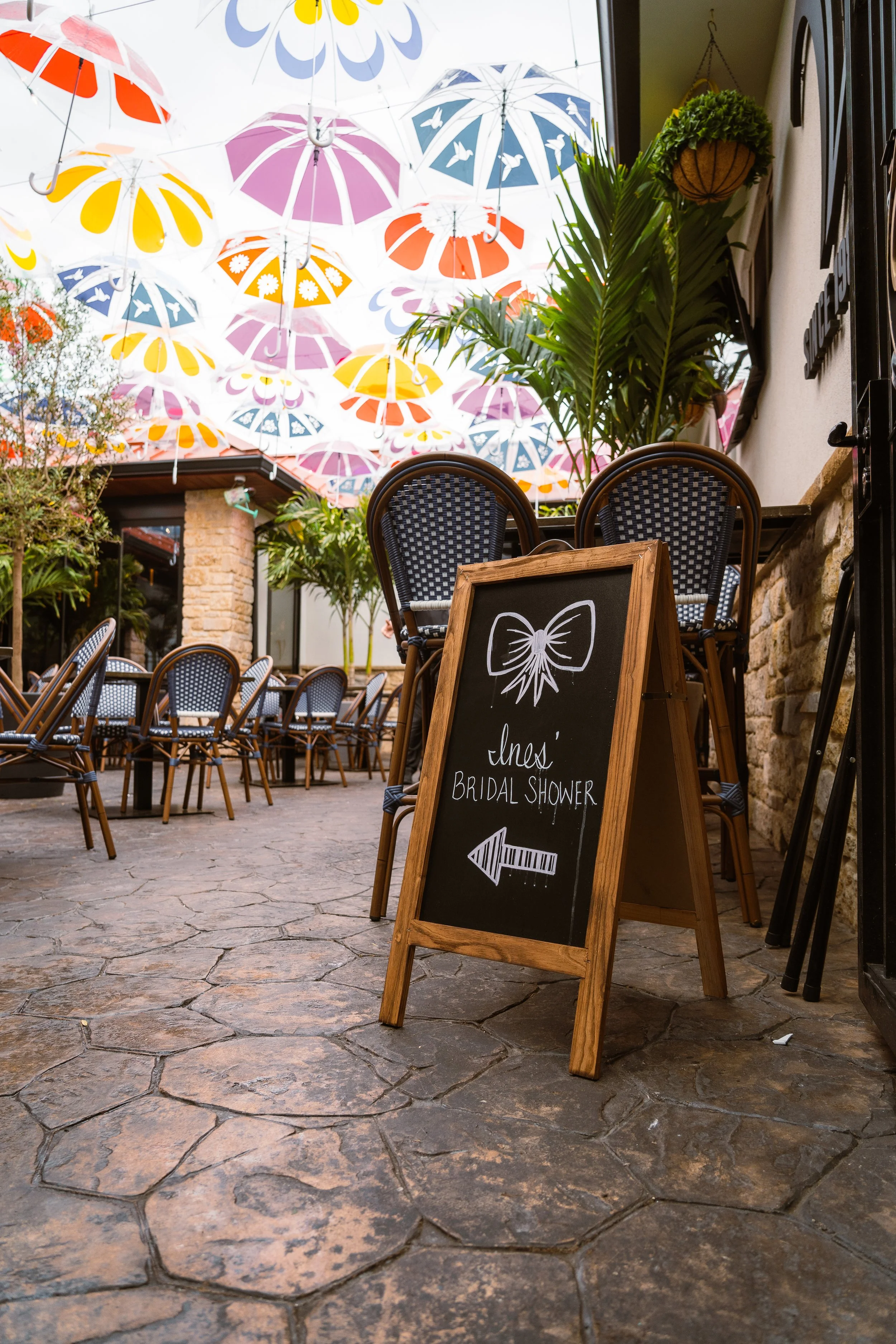 Outdoor patio with tables and chairs beneath colorful umbrellas with a sign for a bridal shower.