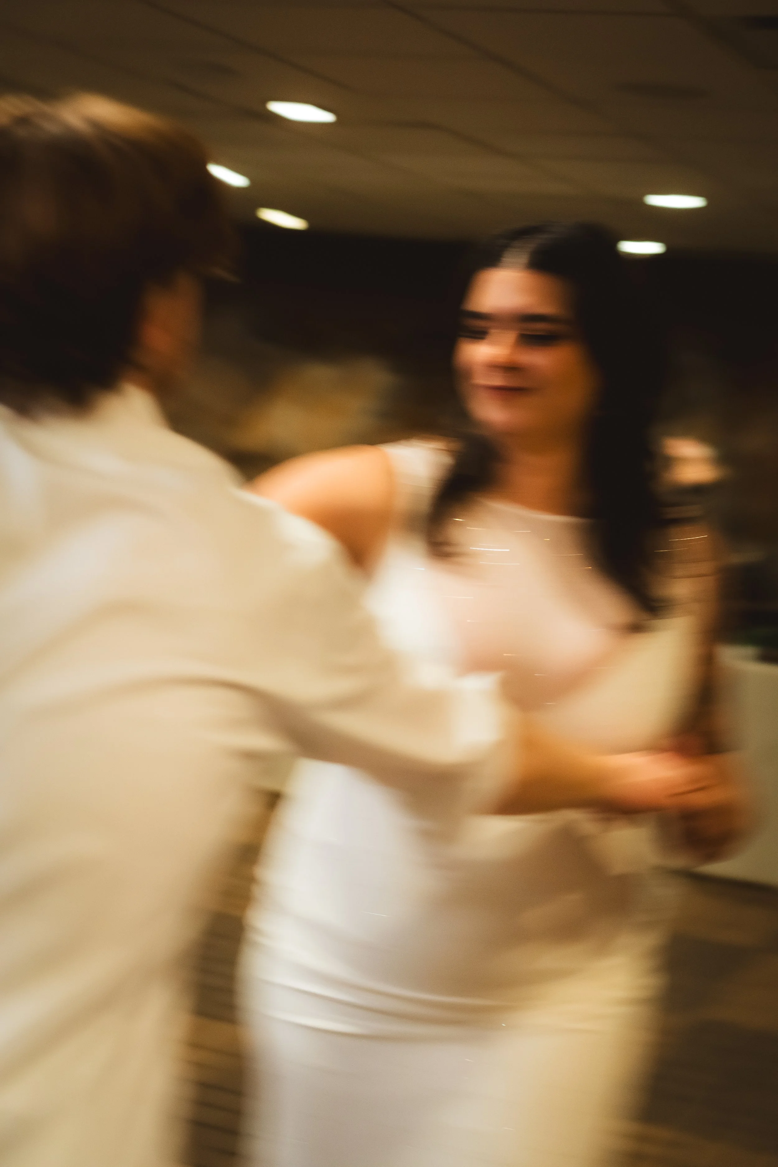 A blurry photo of two women interacting in a dimly lit room, one wearing a white dress with black accents and the other in a white top with a sheer overlay, possibly dancing or engaged in conversation.