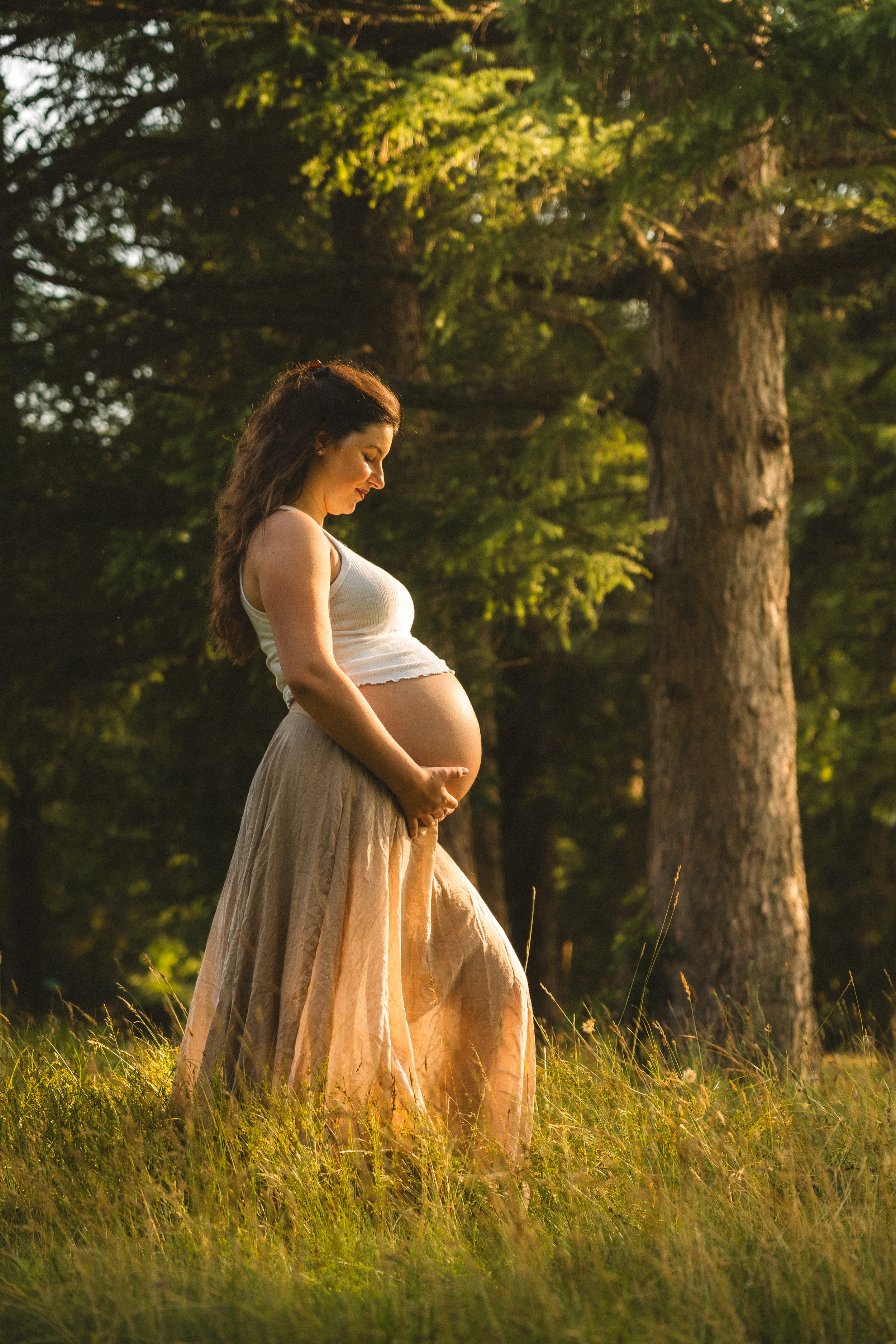 A pregnant woman standing in a grassy field surrounded by trees, gently holding her belly and looking down with a peaceful expression.
