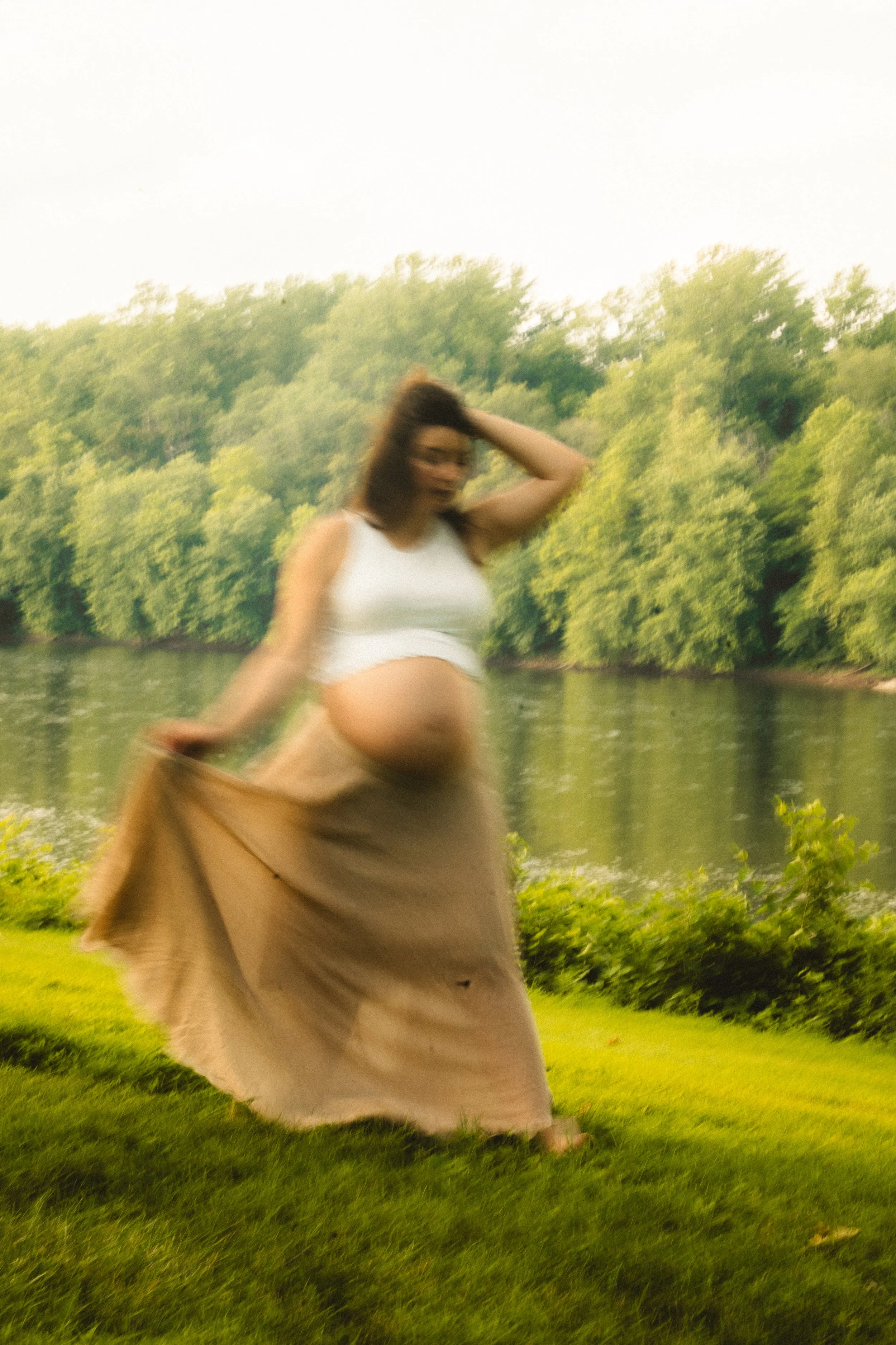A pregnant woman standing outdoors by a river, holding a beige skirt, with trees in the background.