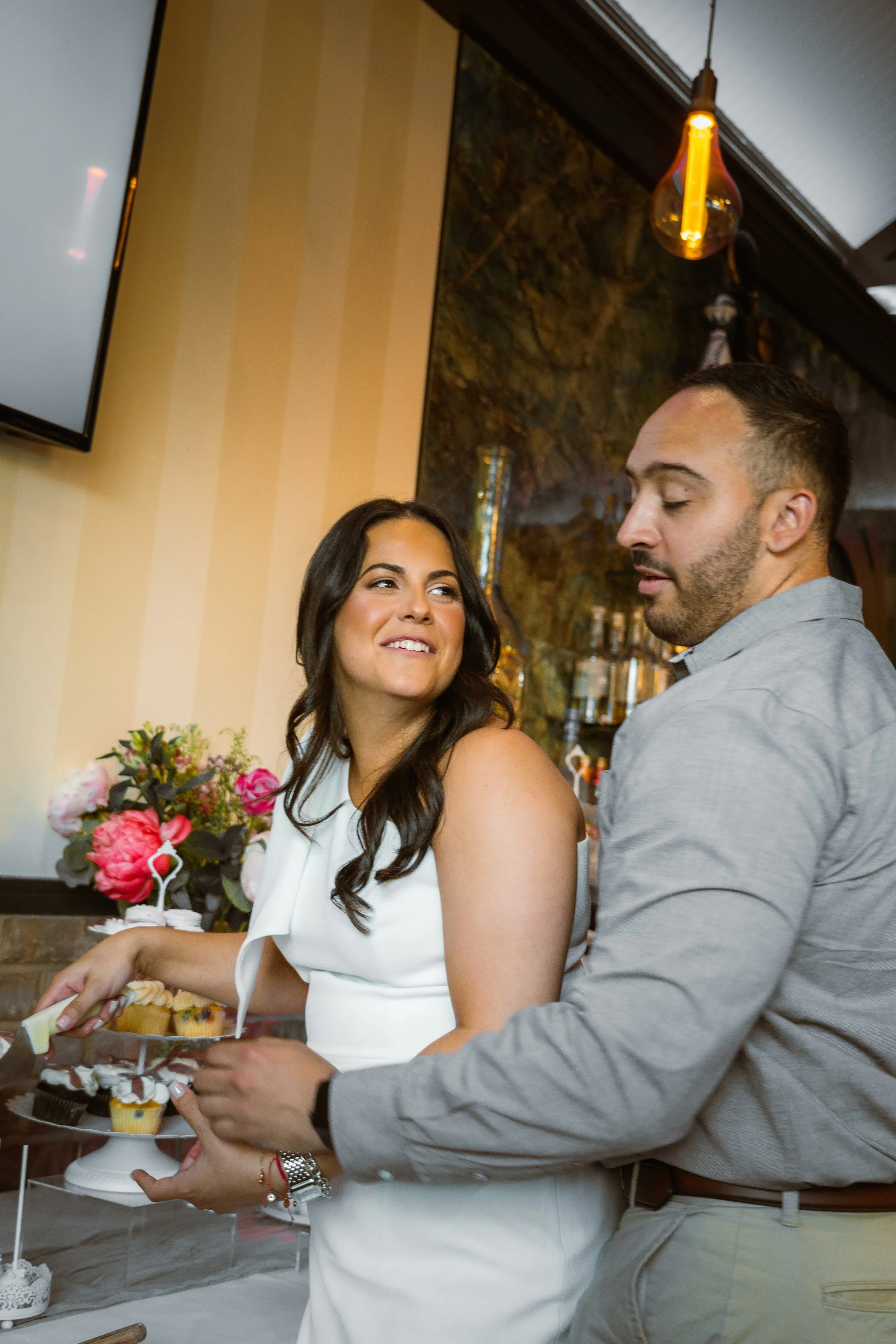 A woman and a man are at a celebration, with the woman smiling while the man offers her a cupcake. There is a floral arrangement and dessert stand in the background.