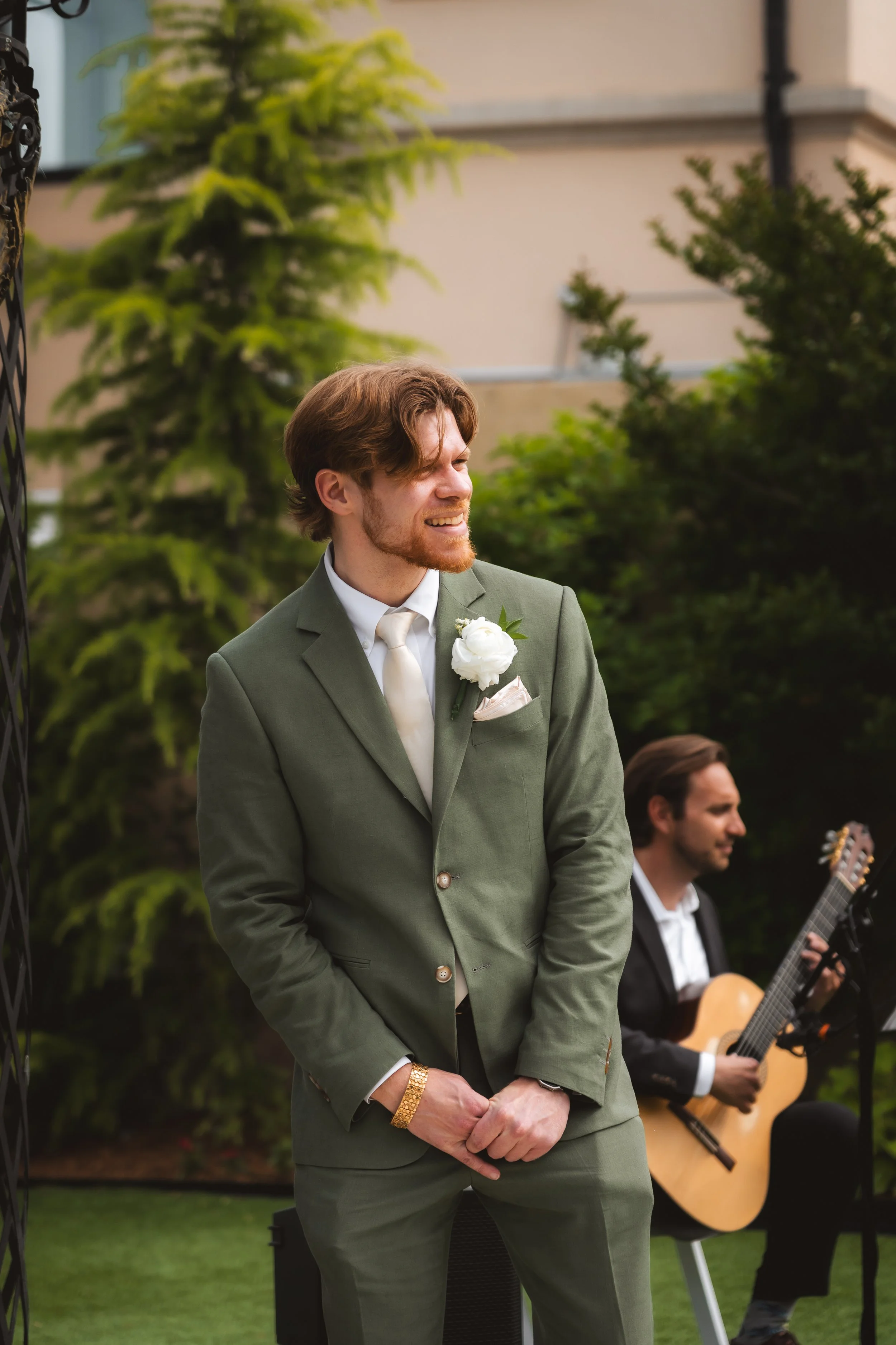 A groom in a green suit stands outdoors during a wedding ceremony, smiling and looking to the side. Behind him, a musician plays guitar, seated on a chair, with lush green trees and the building of a house in the background.