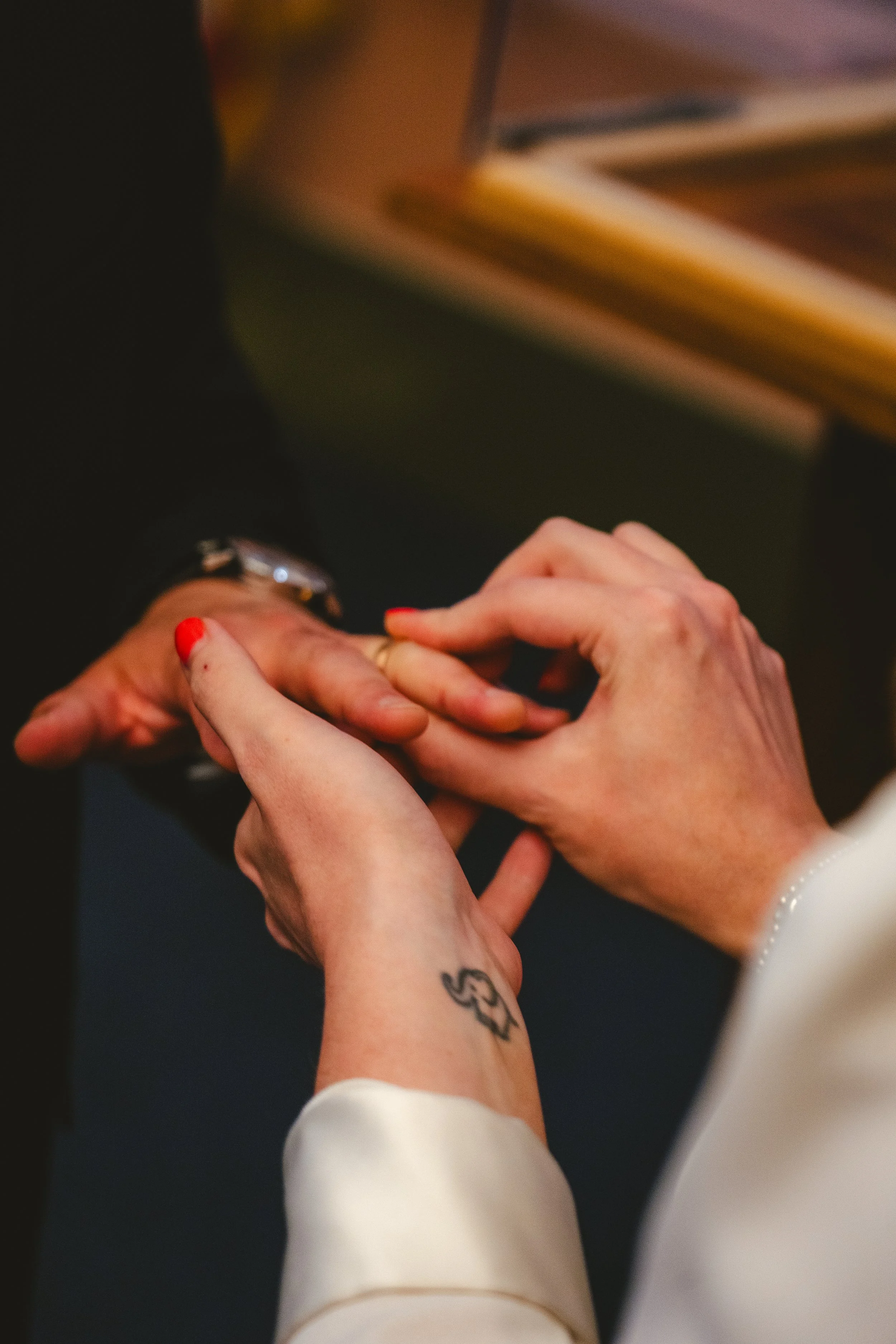 Two people exchanging rings, one person with red nail polish and a tattoo on their wrist. The background includes a desk with a book or binder.