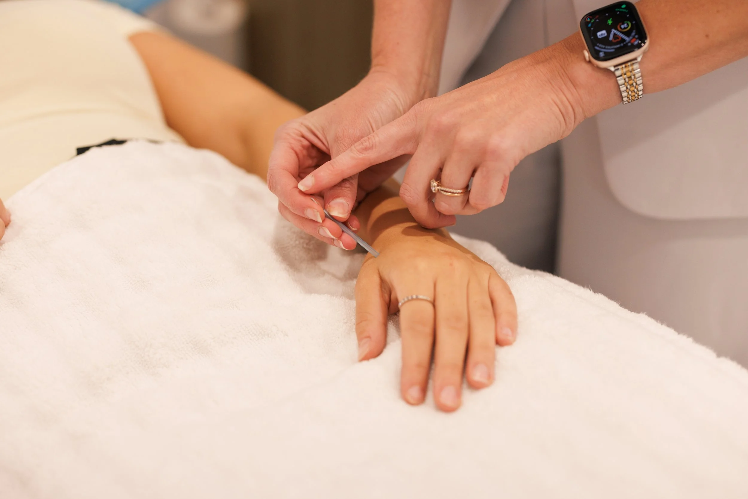 A person receiving a medical injection in their arm by a healthcare worker wearing a smartwatch, with the patient lying on a white towel.