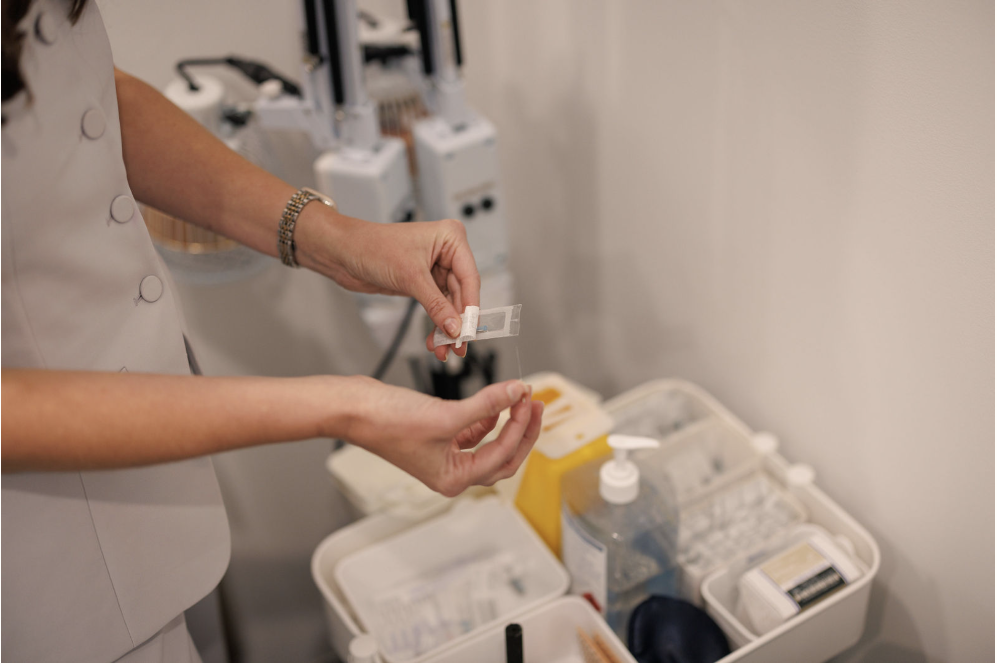 A nurse or healthcare worker preparing a medical or pharmacy injection in a clinical setting, with medical supplies and equipment visible in the background.