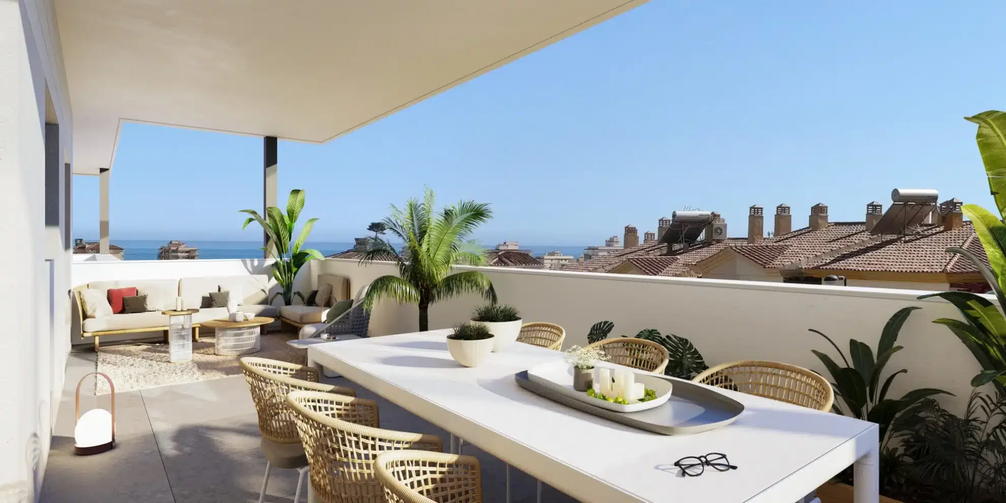 Balcony with outdoor furniture, potted plants, and a view of rooftops and ocean in the distance under a clear blue sky.