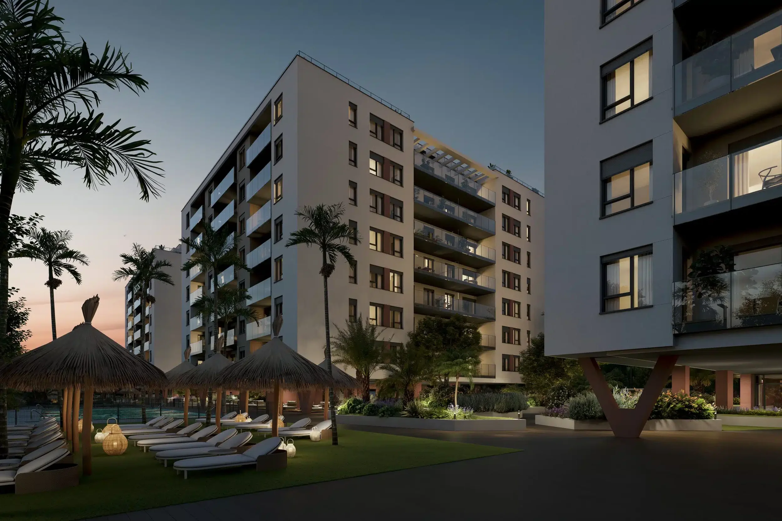 A modern residential complex with multiple apartment buildings, palm trees, and outdoor lounge chairs under thatched umbrellas, illuminated by soft lighting at dusk.
