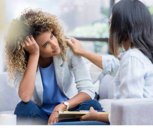 A woman is providing emotional support to a woman in distress during a therapy session in a bright room with large windows.