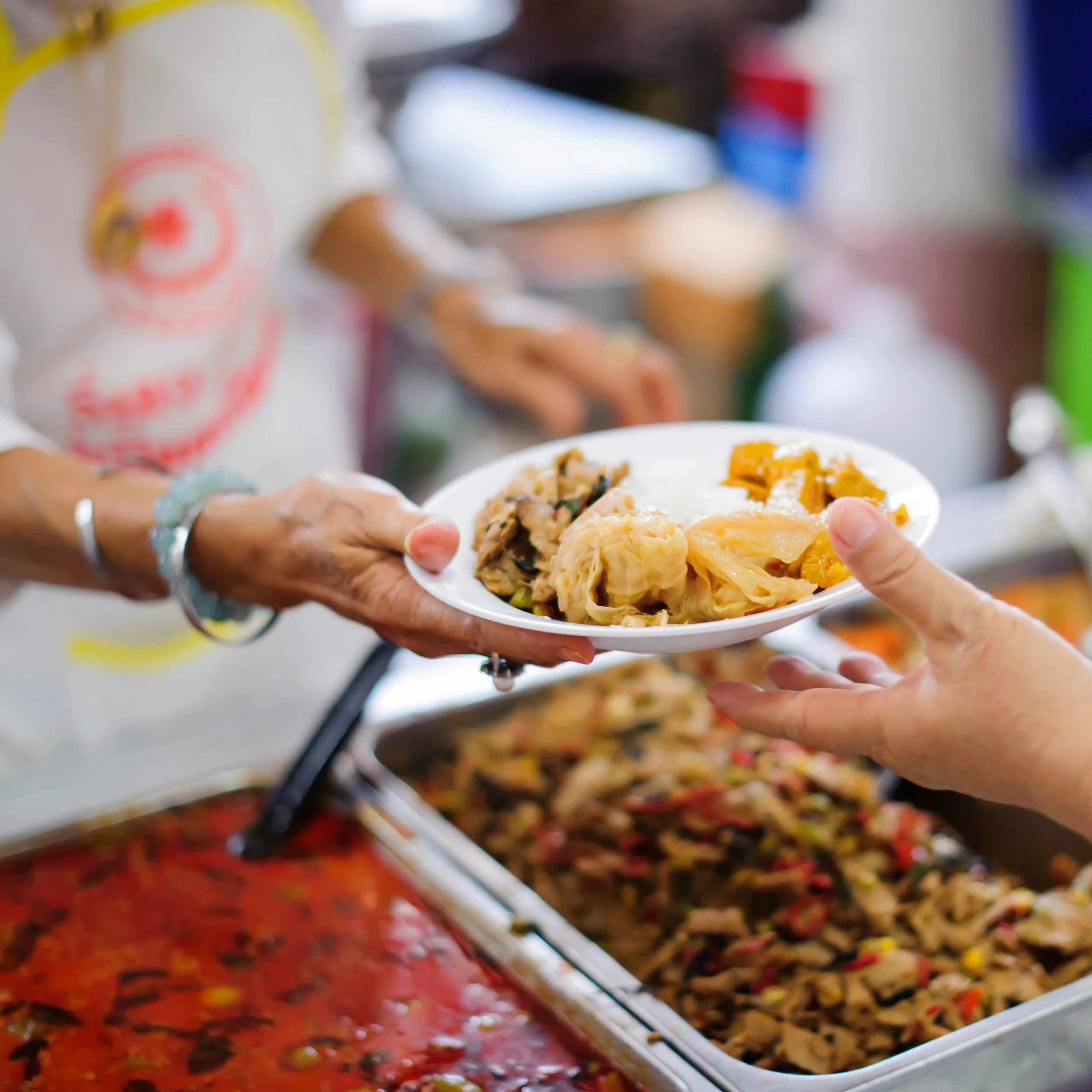 Person handing a plate of food to another person at a buffet with various cooked dishes.