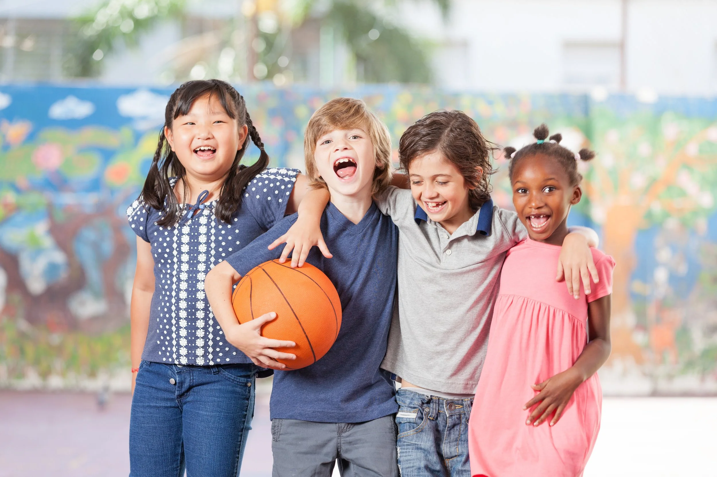 Four children smiling and holding a basketball together, standing closely with arms around each other in an indoor play area with colorful murals in the background.