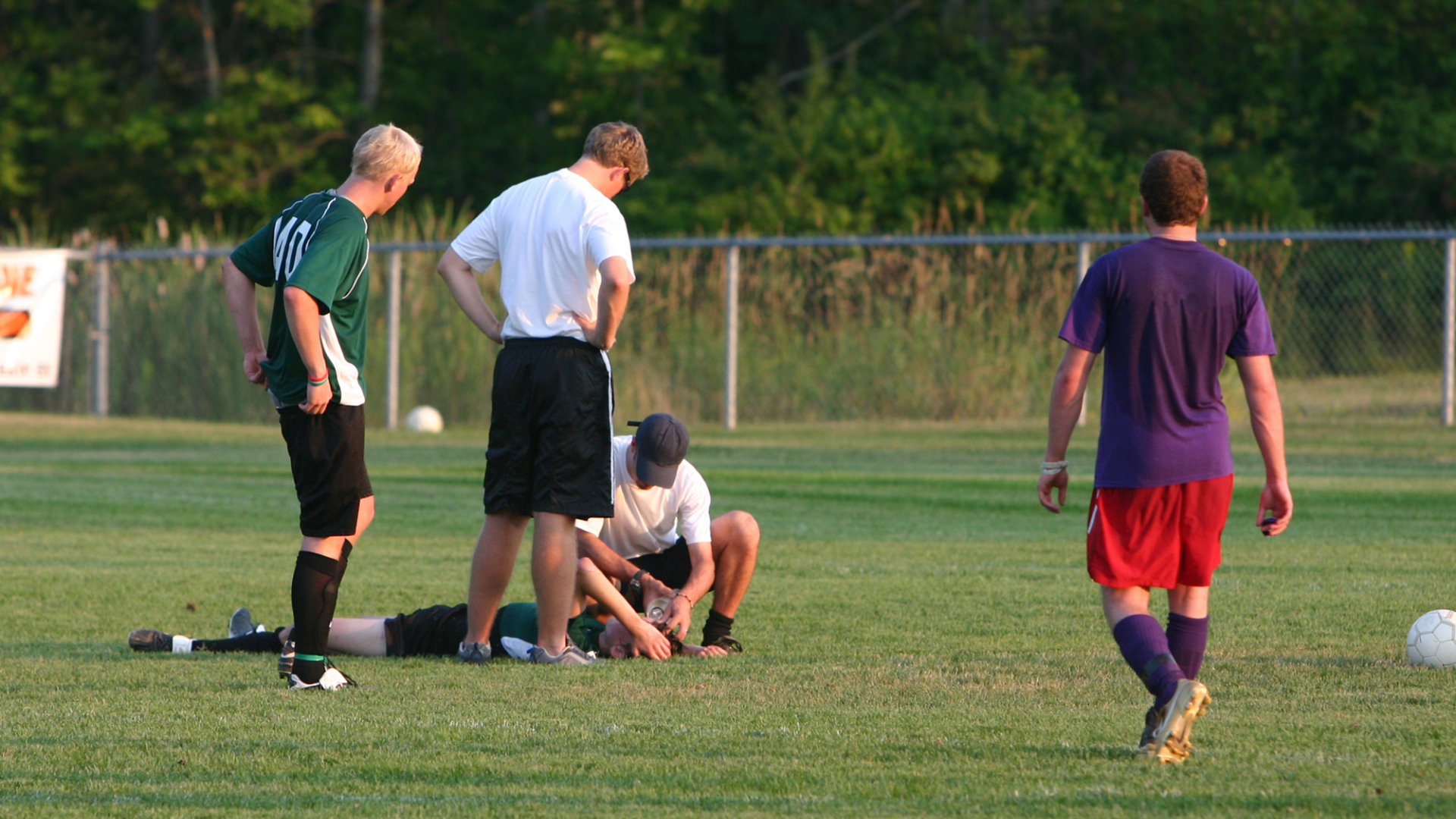 A group of young men are on a soccer field; one is lying on the grass with a head injury, others are attending to him, and a few are standing nearby.