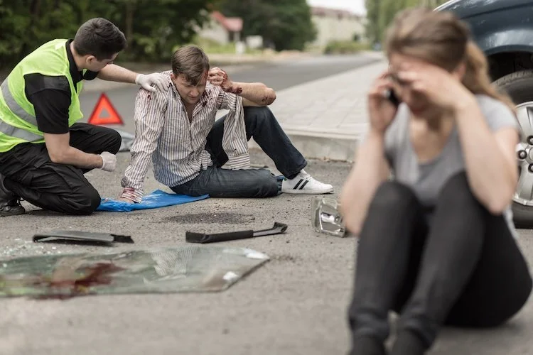 A person injured on the roadside being attended to by paramedics, with a woman distressed talking on the phone nearby.