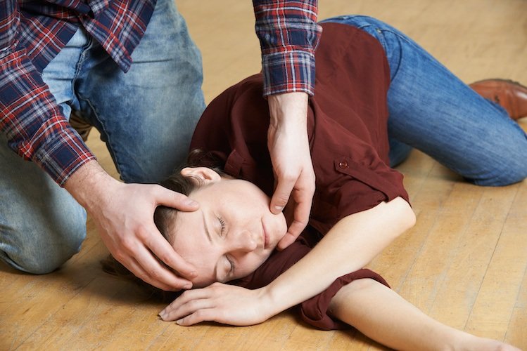 A person performing CPR on a young child lying on the floor with eyes closed, wearing a maroon shirt and blue jeans.
