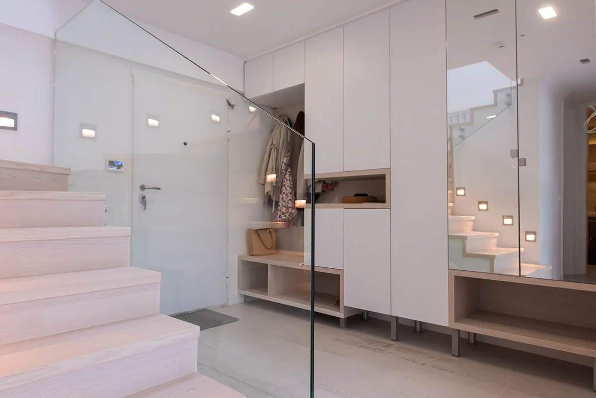 A modern, minimalist entryway with white cabinetry, a glass partition, and stairs with built-in lighting.
