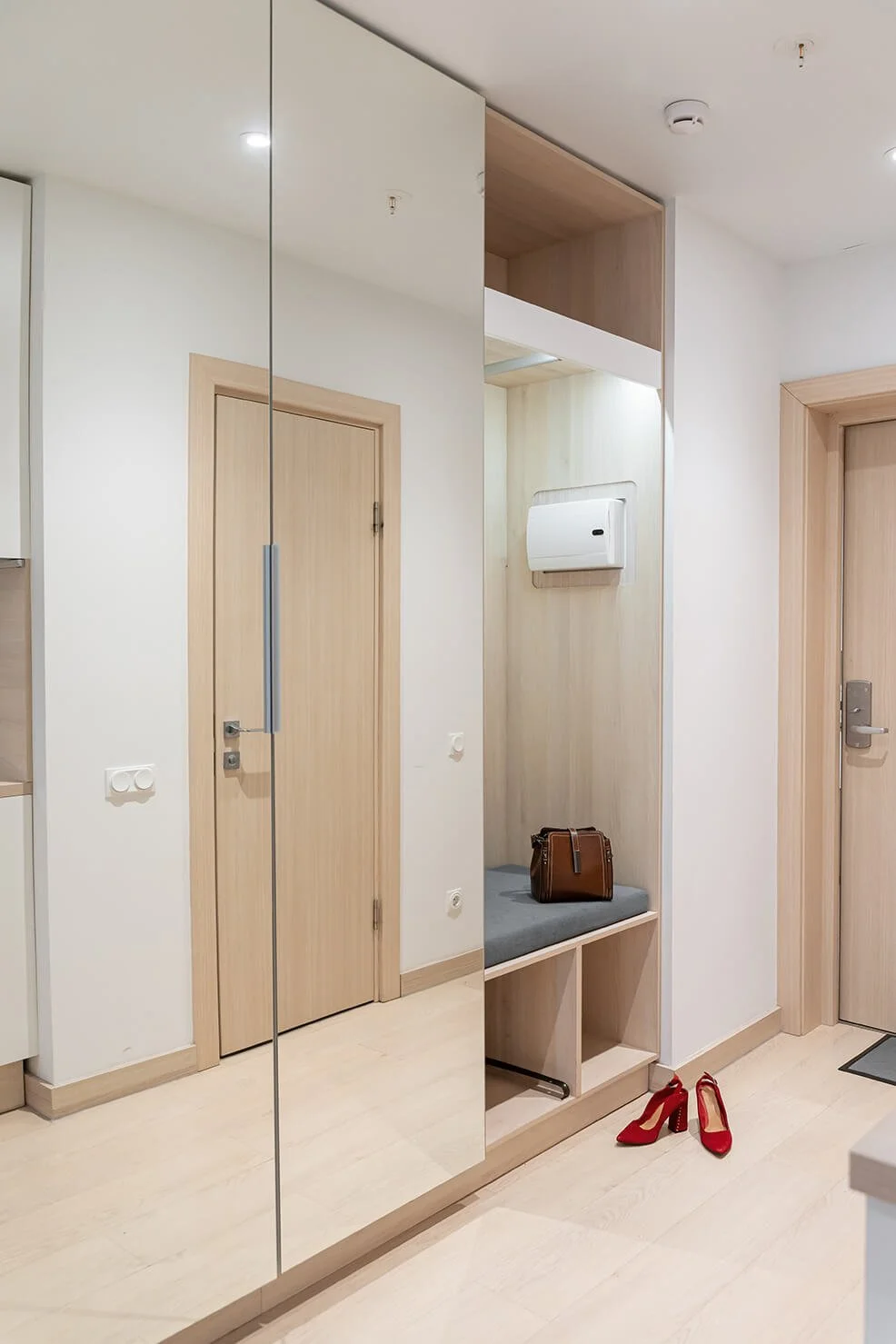A modern entryway with light wood doors, a bench with a gray cushion, a brown handbag, a pair of red high-heeled shoes, and a full-length mirror.