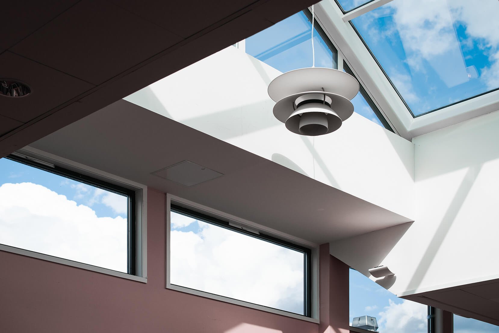 Interior view of a modern building with large windows and skylights, showing a white ceiling with a contemporary pendant light fixture and a blue sky with clouds outside.