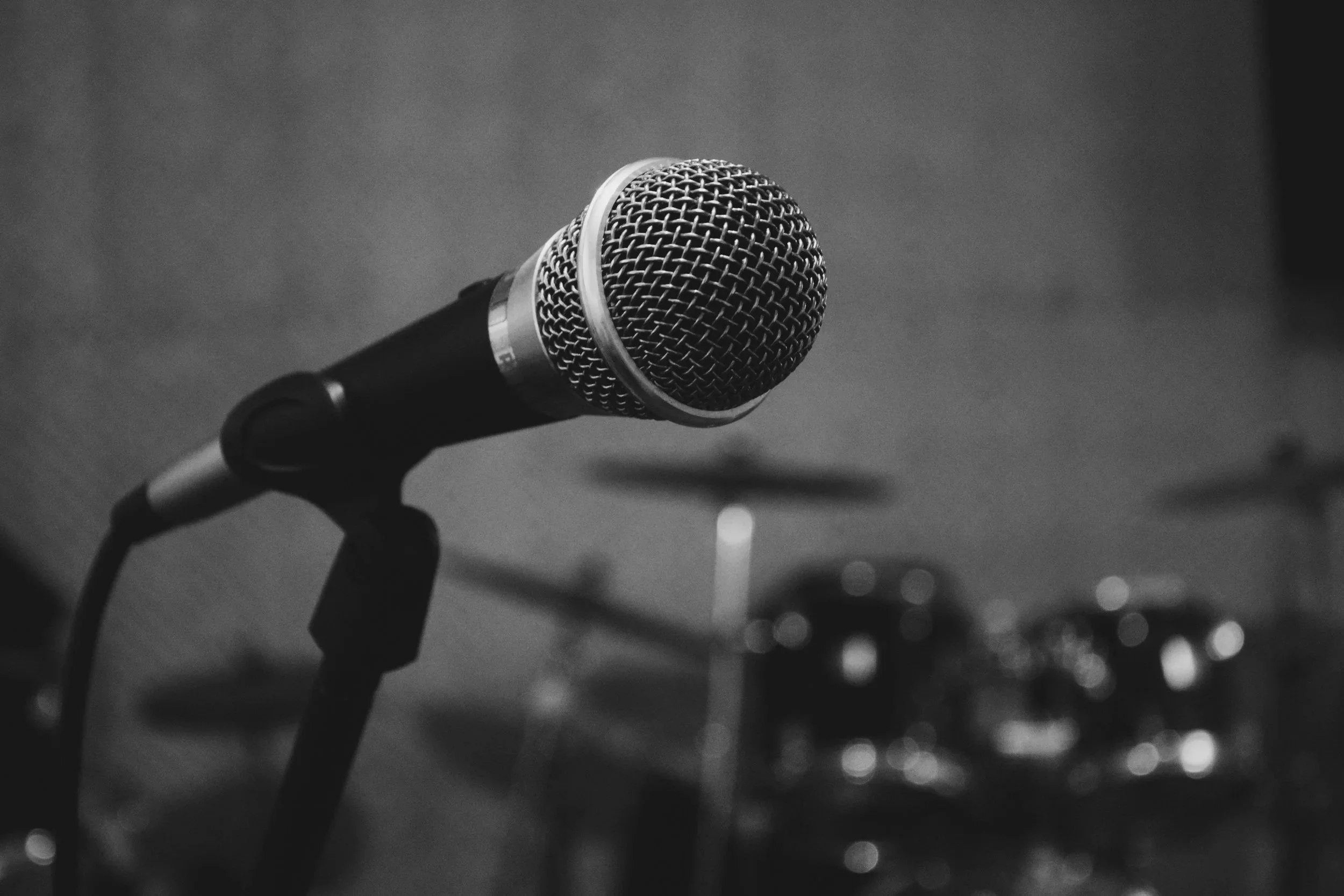 Close-up of a microphone on a stand, with drums in the blurry background.