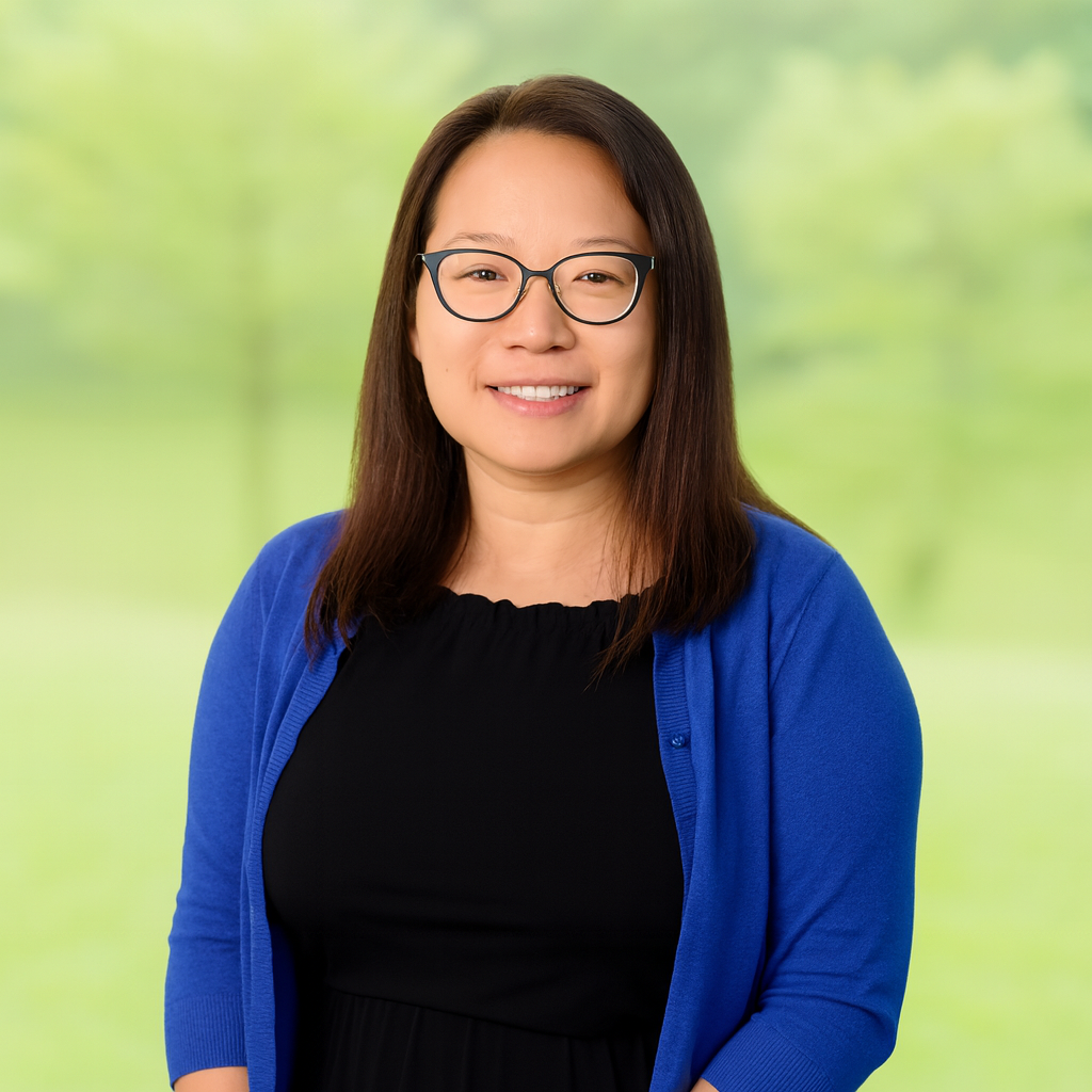 A woman with shoulder-length dark hair, glasses, and a black top wearing a blue cardigan, smiling outdoors with a blurred green background.