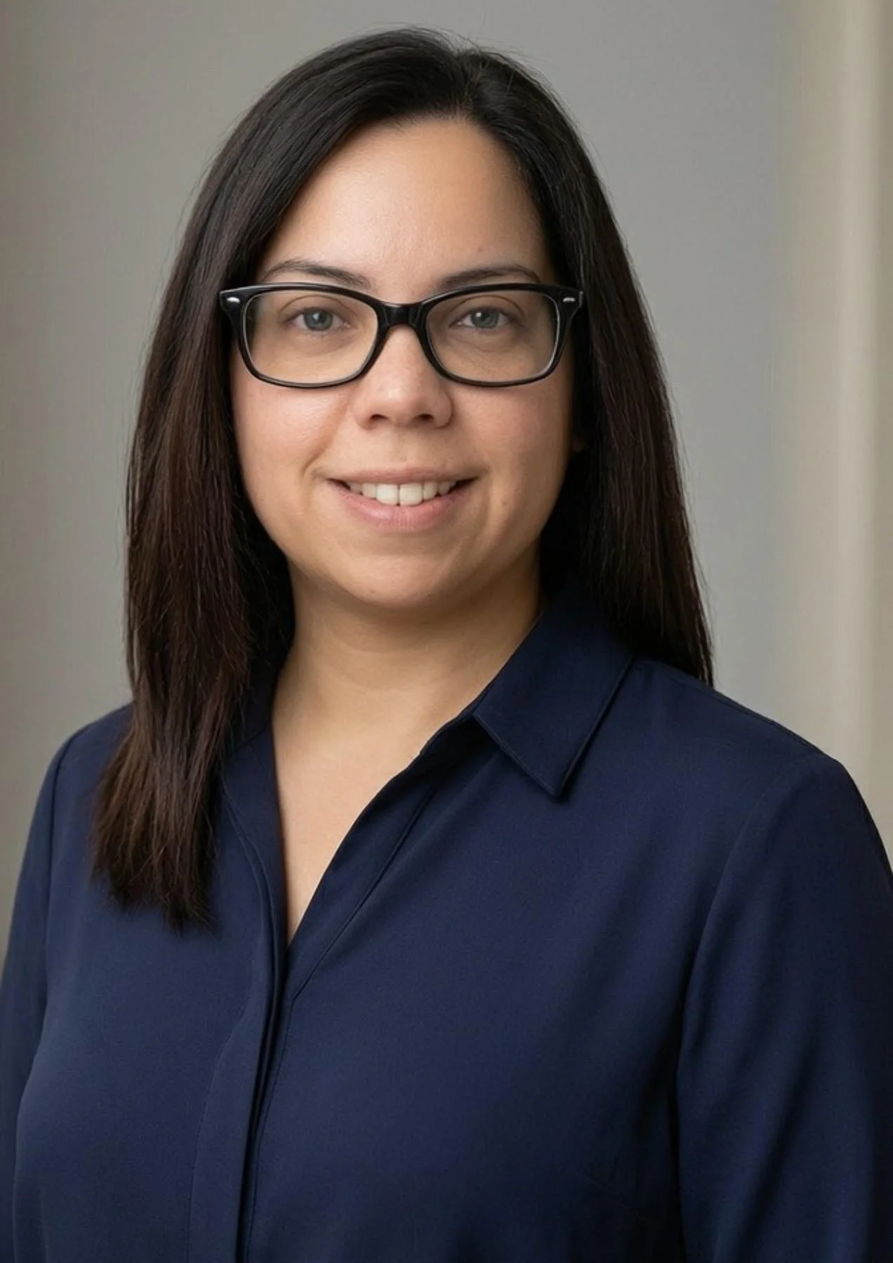Portrait of a woman with straight dark brown hair, glasses, light skin, wearing a navy blue blouse, smiling slightly, against a neutral background.