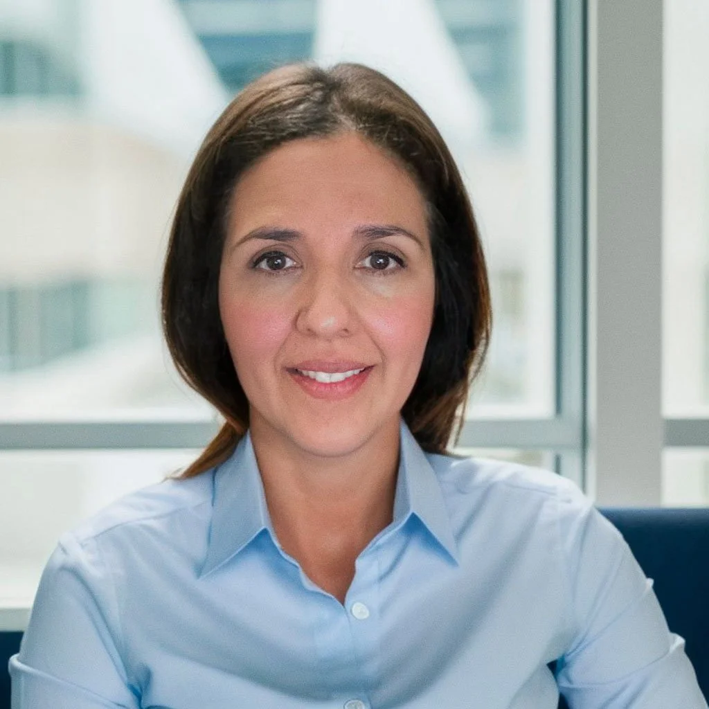 A woman with shoulder-length brown hair smiling while sitting in front of a window with a cityscape view in the background.