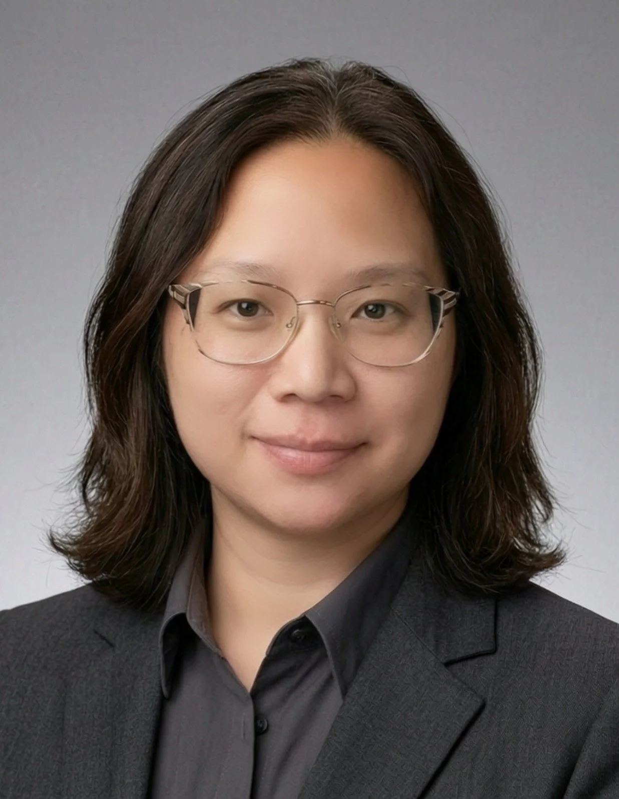Professional portrait of a woman with shoulder-length dark hair, wearing glasses and a dark blazer over a gray shirt, against a plain gray background.