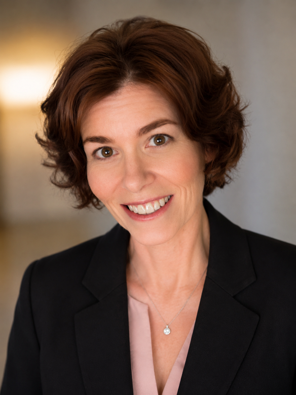 A smiling woman with short brown curly hair wearing a black blazer and a light pink blouse, smiling at the camera.