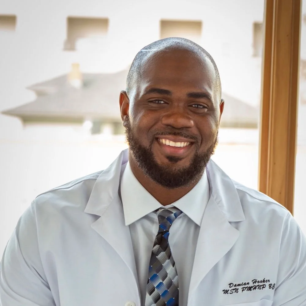 A smiling man in a white medical coat and patterned tie, sitting indoors near a window.