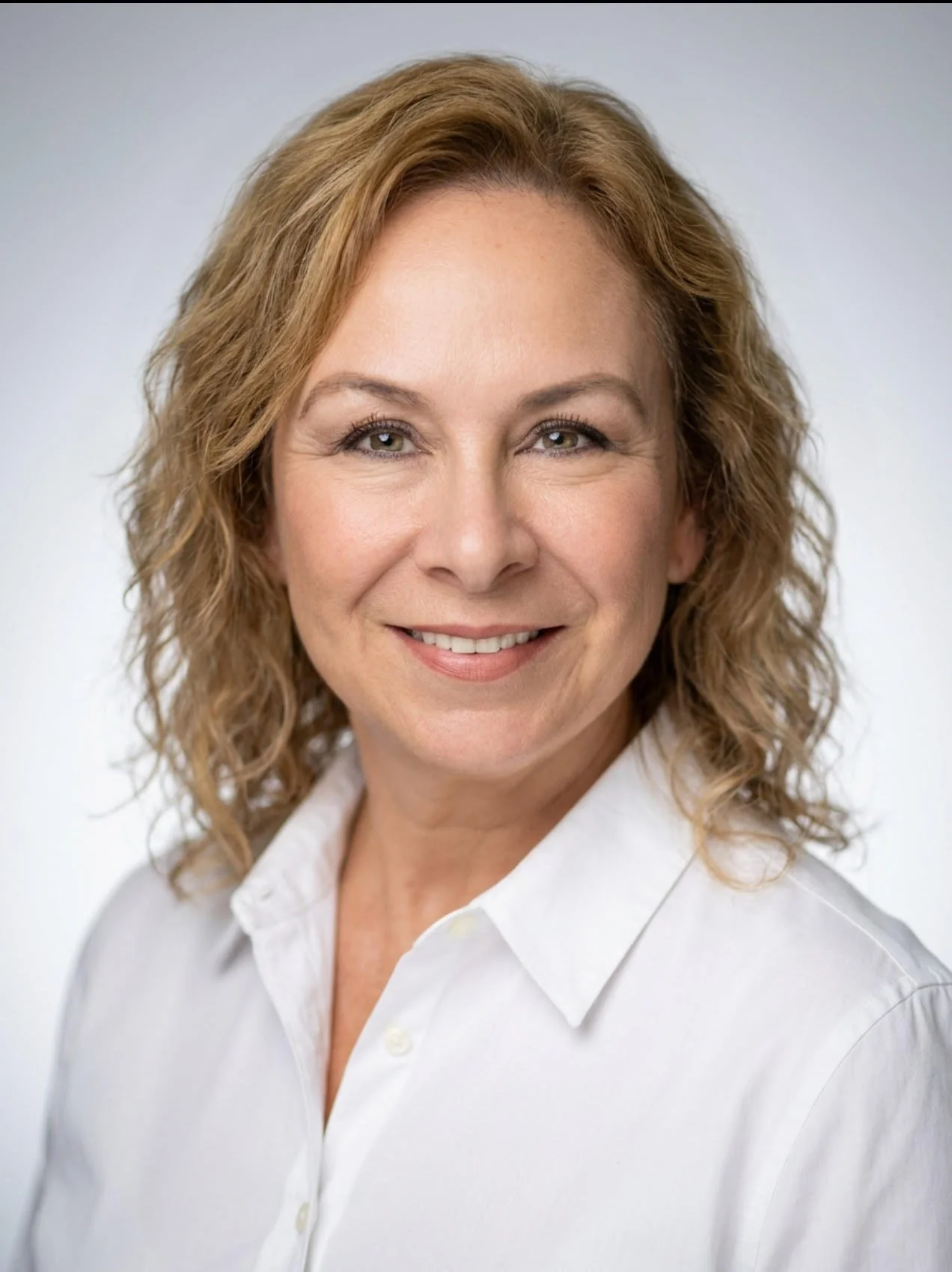A woman with wavy blonde hair wearing a white shirt and smiling in front of a plain white background.
