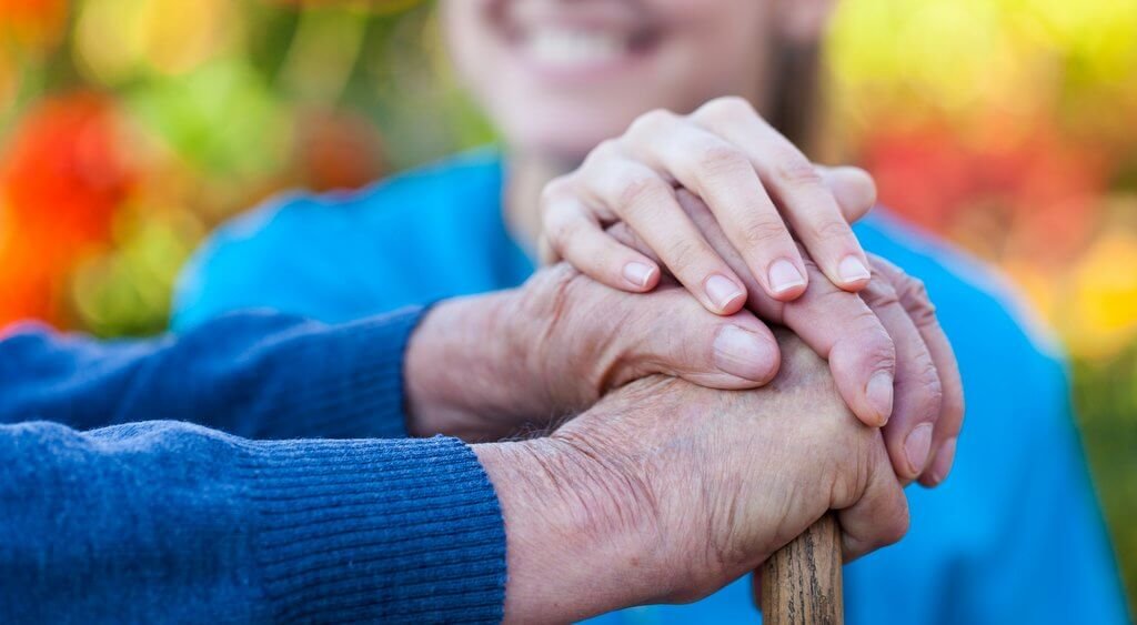 Close-up of two people holding hands, one elderly and one younger, outdoors with colorful blurred background.