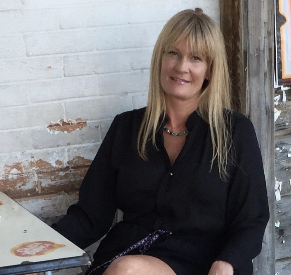 A woman with blonde hair, wearing a black shirt and a colorful necklace, sitting outdoors near a white brick wall and a wooden structure.