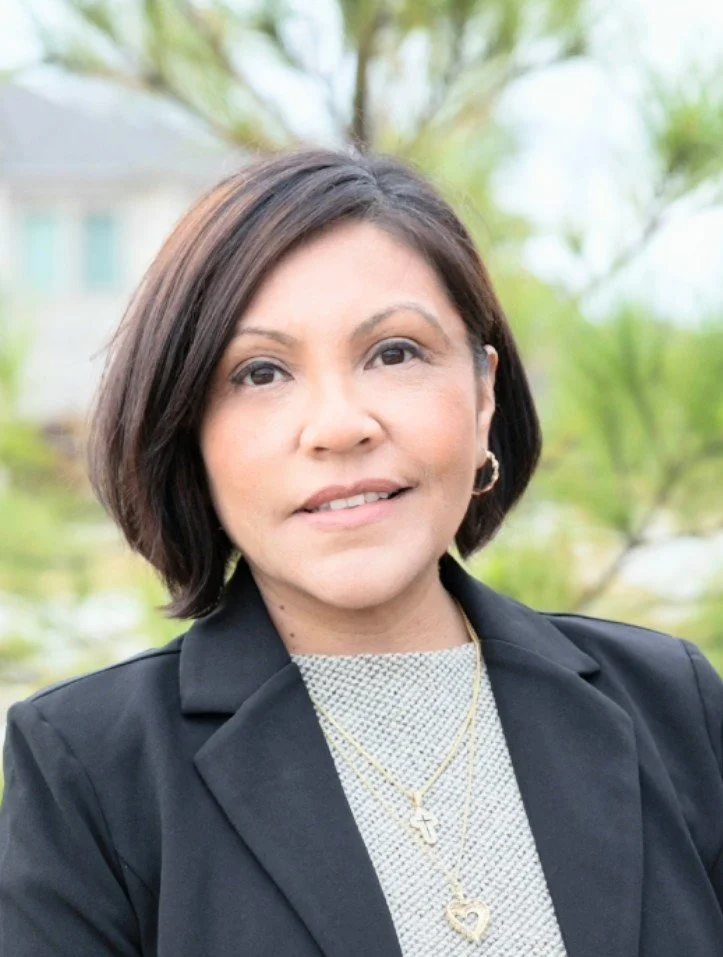 A woman with short brown hair wearing a black blazer, layered necklaces including a cross and heart, and small hoop earrings, outdoors with green trees and a house in the background.